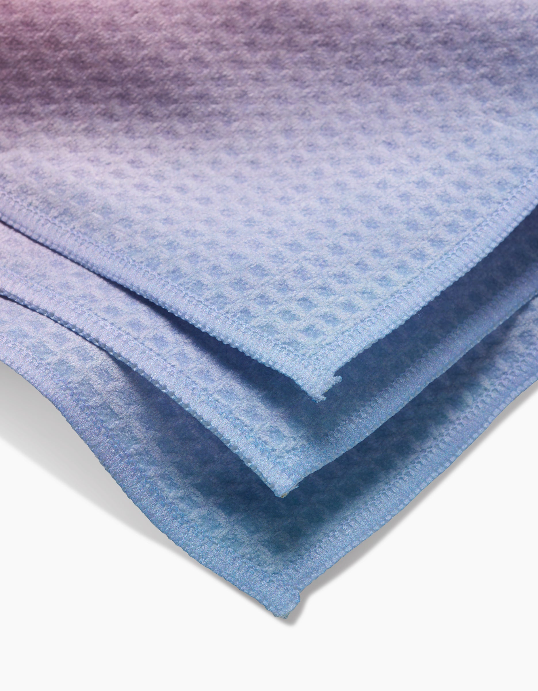A close-up of neatly folded, light blue GEOMETRY Aura washcloths highlights their Geoweave technology with a textured, waffle-like pattern, soft material, and clean edges.