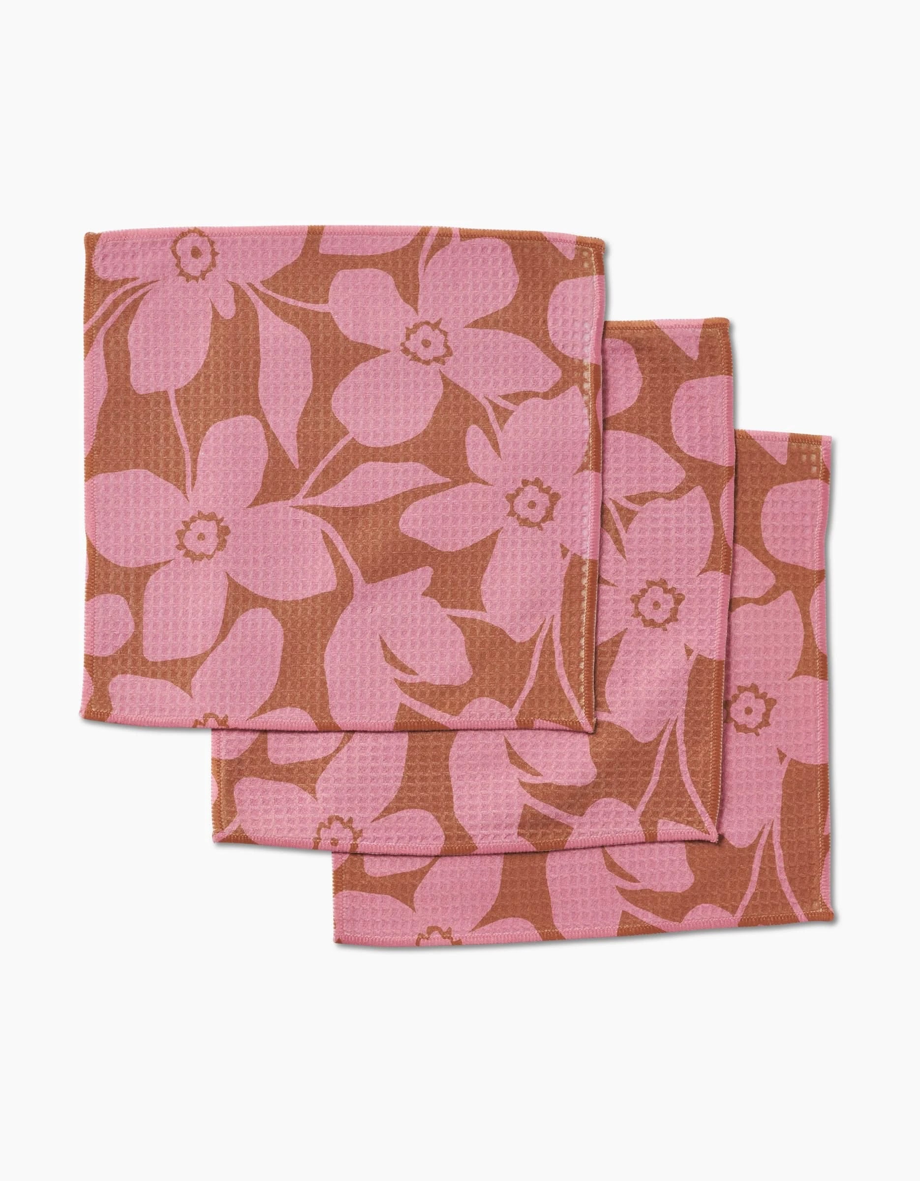 Three Big Fuchsia Flowers cloth napkins by GEOMETRY, featuring a pink floral design on brown and made with ultra-absorbent Geoweave technology, are shown stacked in a staggered arrangement on a white surface.