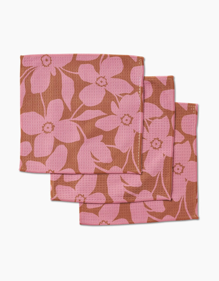 Three Big Fuchsia Flowers cloth napkins by GEOMETRY, featuring a pink floral design on brown and made with ultra-absorbent Geoweave technology, are shown stacked in a staggered arrangement on a white surface.