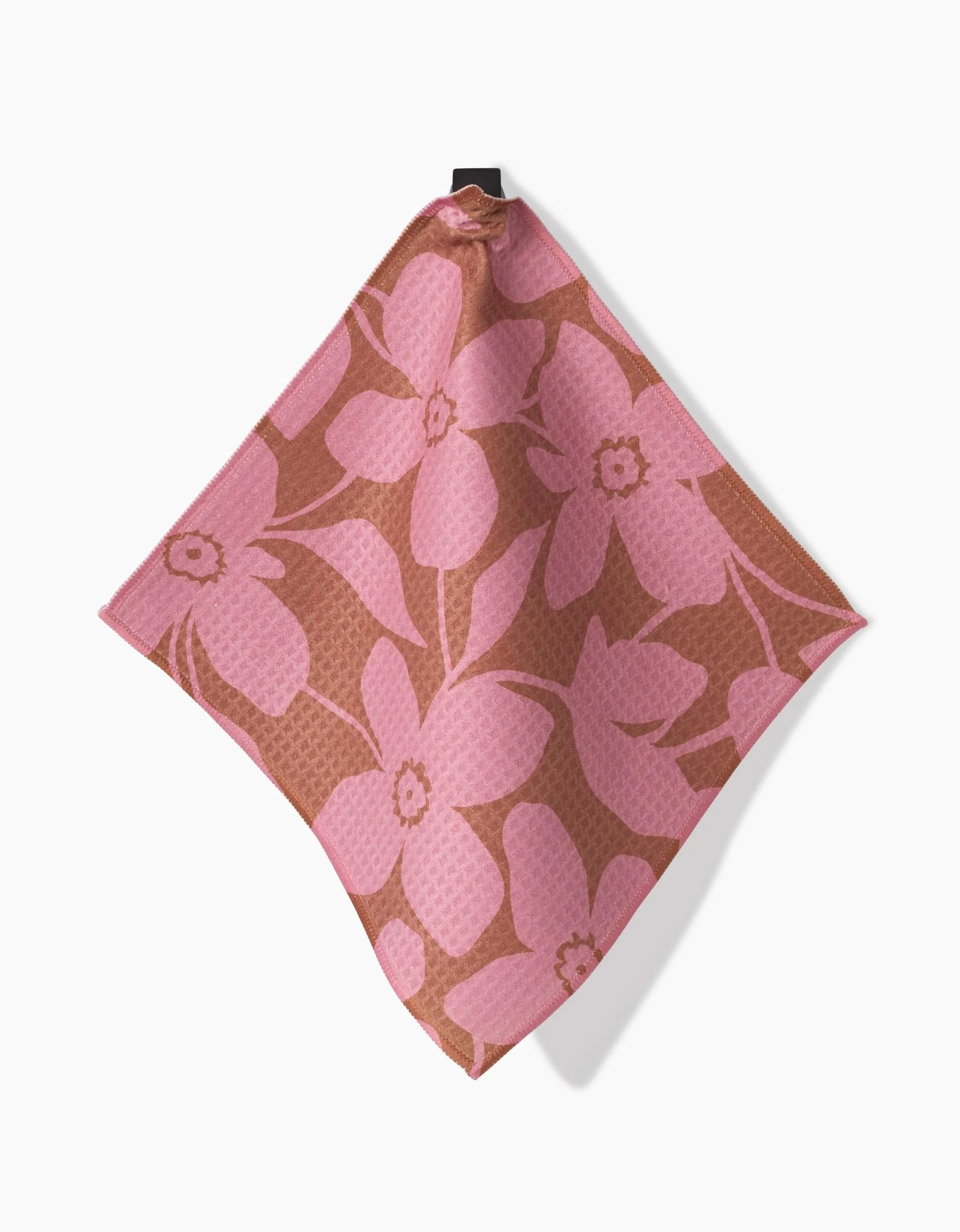 The GEOMETRY Big Fuchsia Flowers Luxe Washcloth features a pink floral design on brown, made with ultra-absorbent Geoweave technology, and is shown hanging from one corner against a white background.