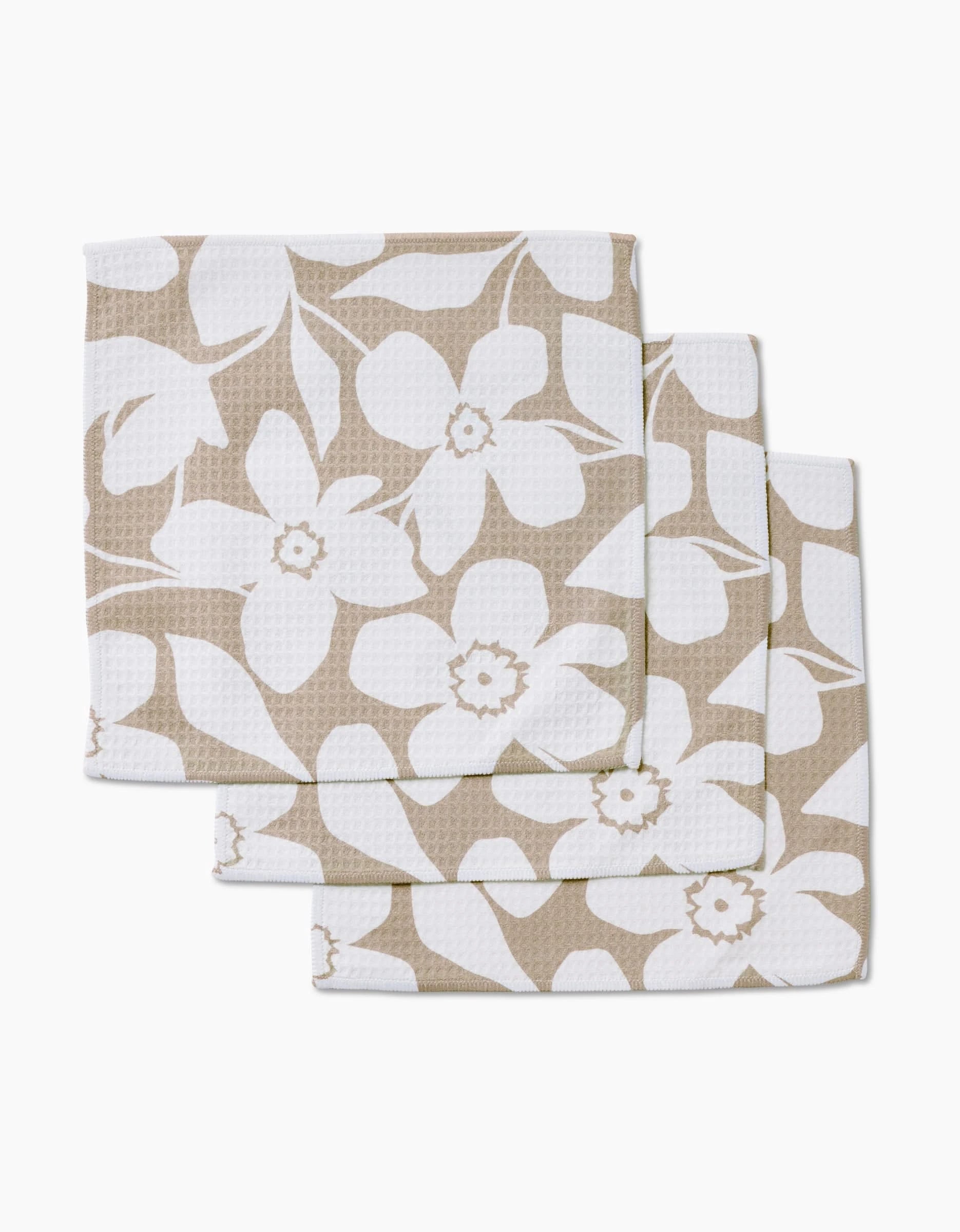 Three stacked GEOMETRY Big Tan Flowers beige cloth napkins, featuring a white floral pattern of large flowers and leaves, made with ultra-absorbent Geoweave technology, displayed on a white background.