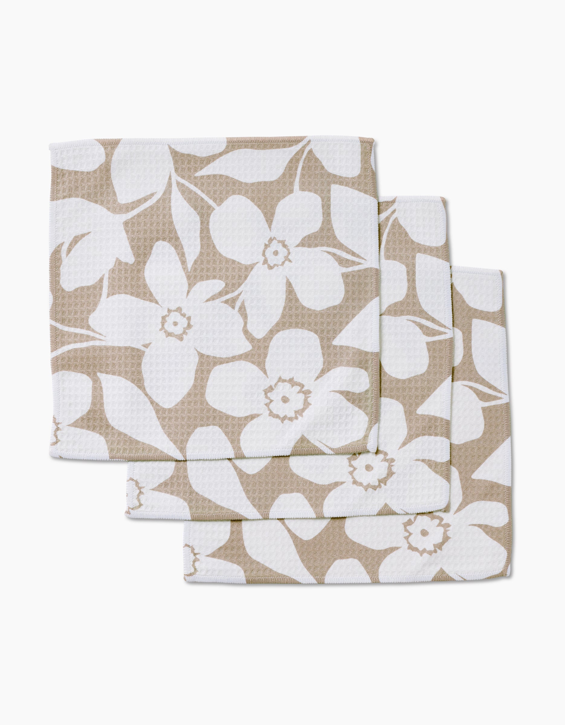 Three stacked GEOMETRY Big Tan Flowers beige cloth napkins, featuring a white floral pattern of large flowers and leaves, made with ultra-absorbent Geoweave technology, displayed on a white background.
