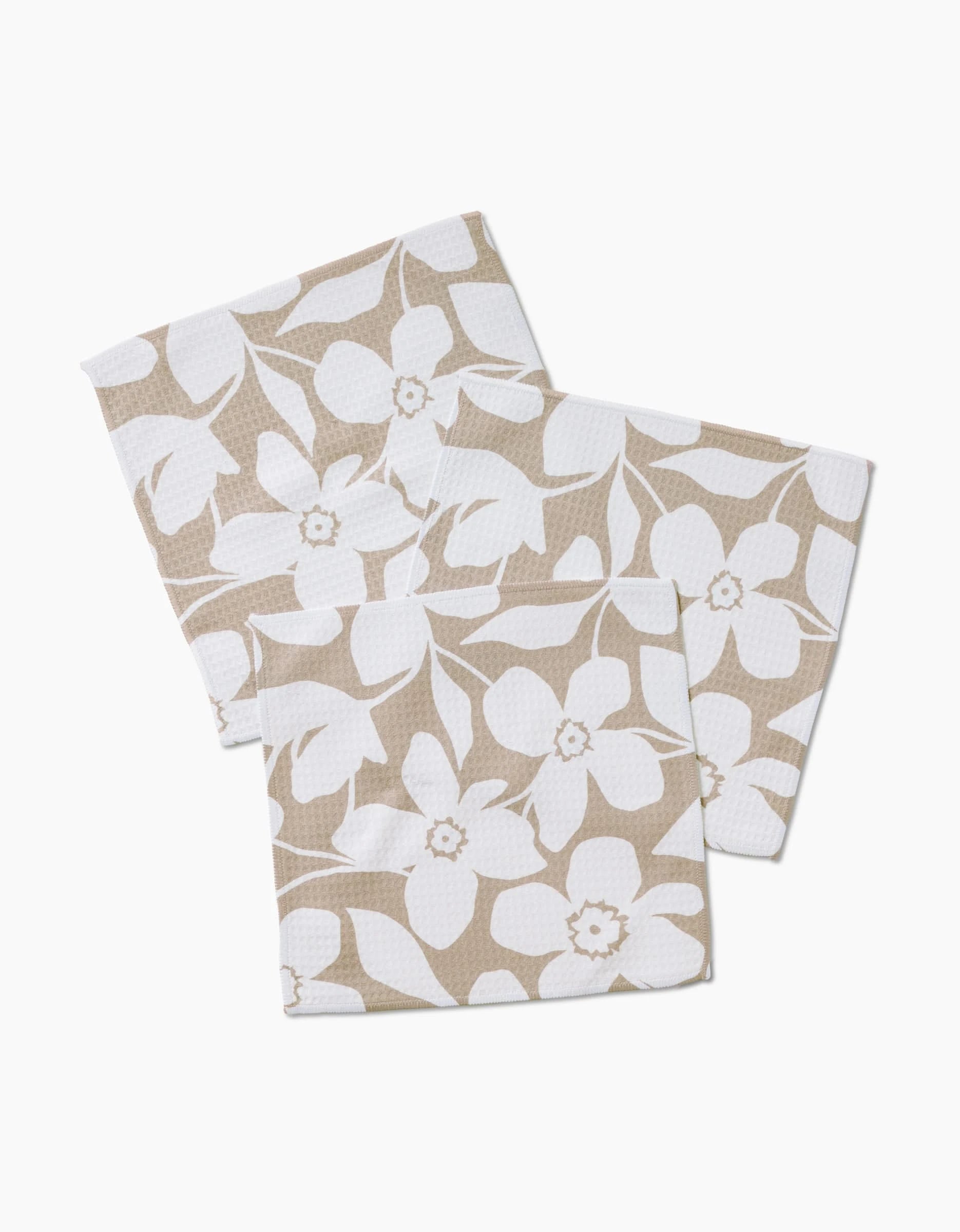 Three GEOMETRY Big Tan Flowers washcloths, featuring a beige hue with white floral patterns and Geoweave technology, are arranged overlapping each other on a white background.