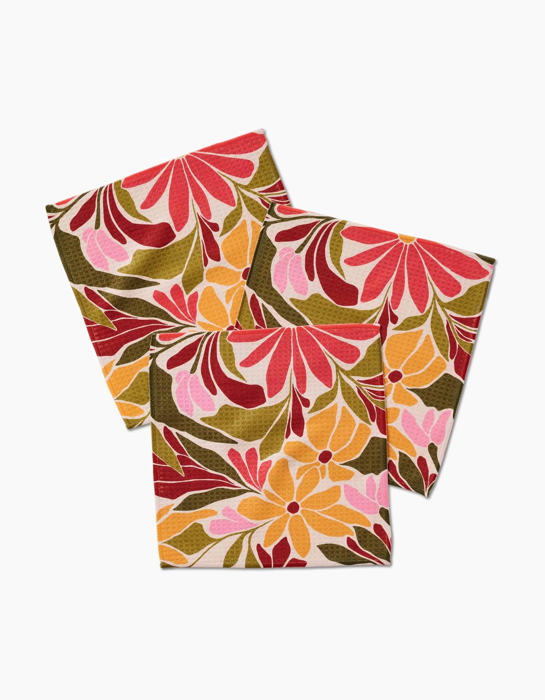 Three Blooming Tan napkins by GEOMETRY, featuring a vibrant floral pattern in red, pink, yellow, and green on recycled fabric, are arranged overlapping on a white background.