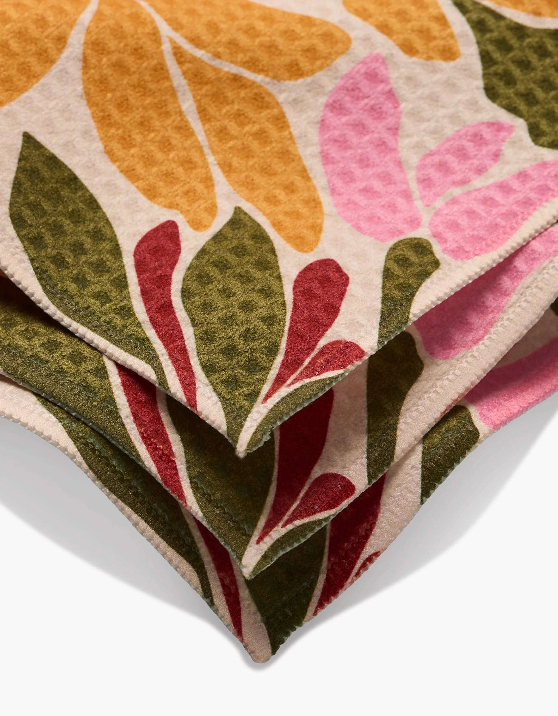 A close-up of overlapping GEOMETRY Blooming Tan washcloths with a textured surface, showcasing a vibrant leaf pattern in yellow, pink, green, and red on a light background.