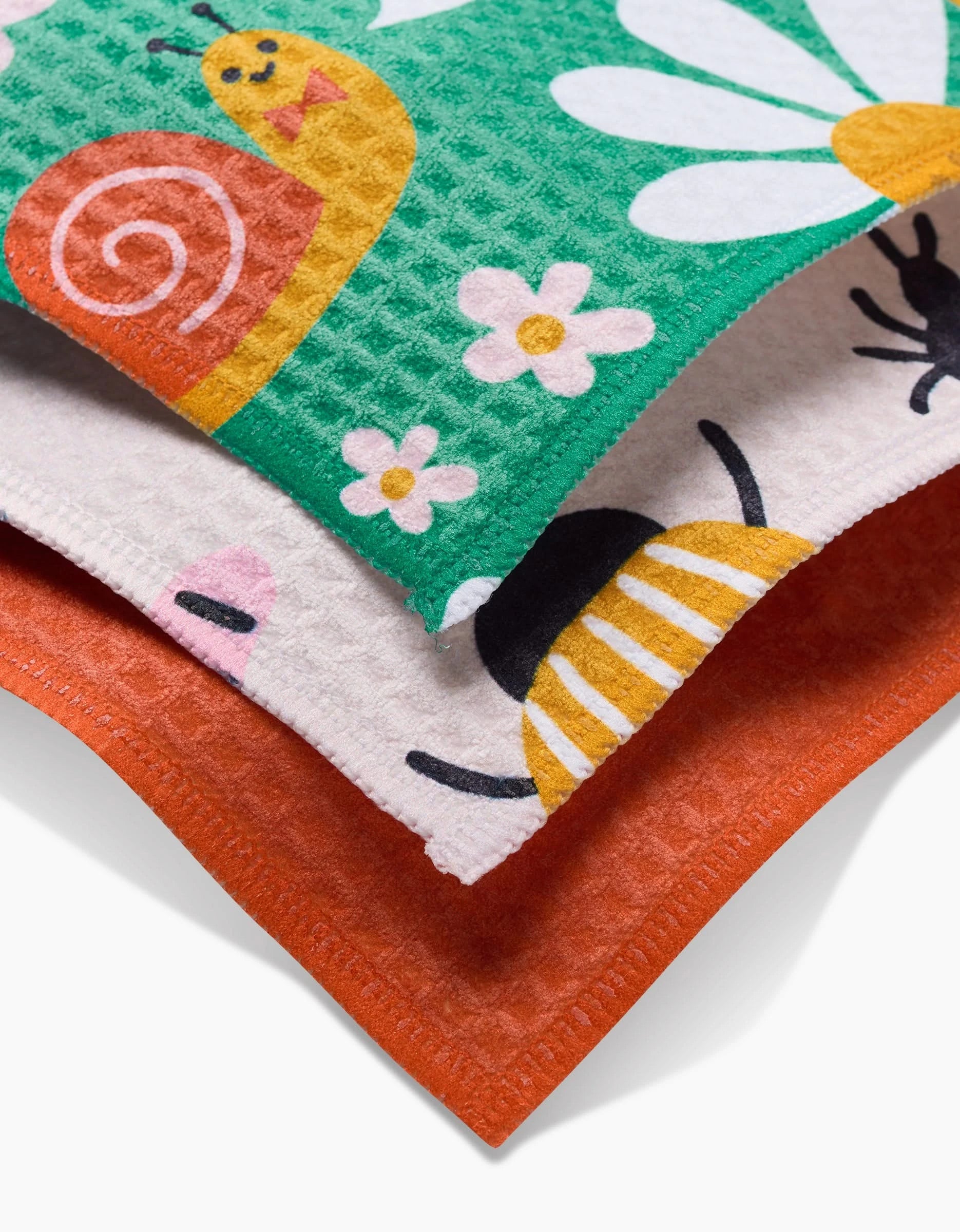 The Bugs dishcloth set by Genna Blackburn features three ultra-absorbent cloths: one with a smiling snail and daisies, one with part of a bee design, and a solid orange. The textured surface ensures effortless cleaning.