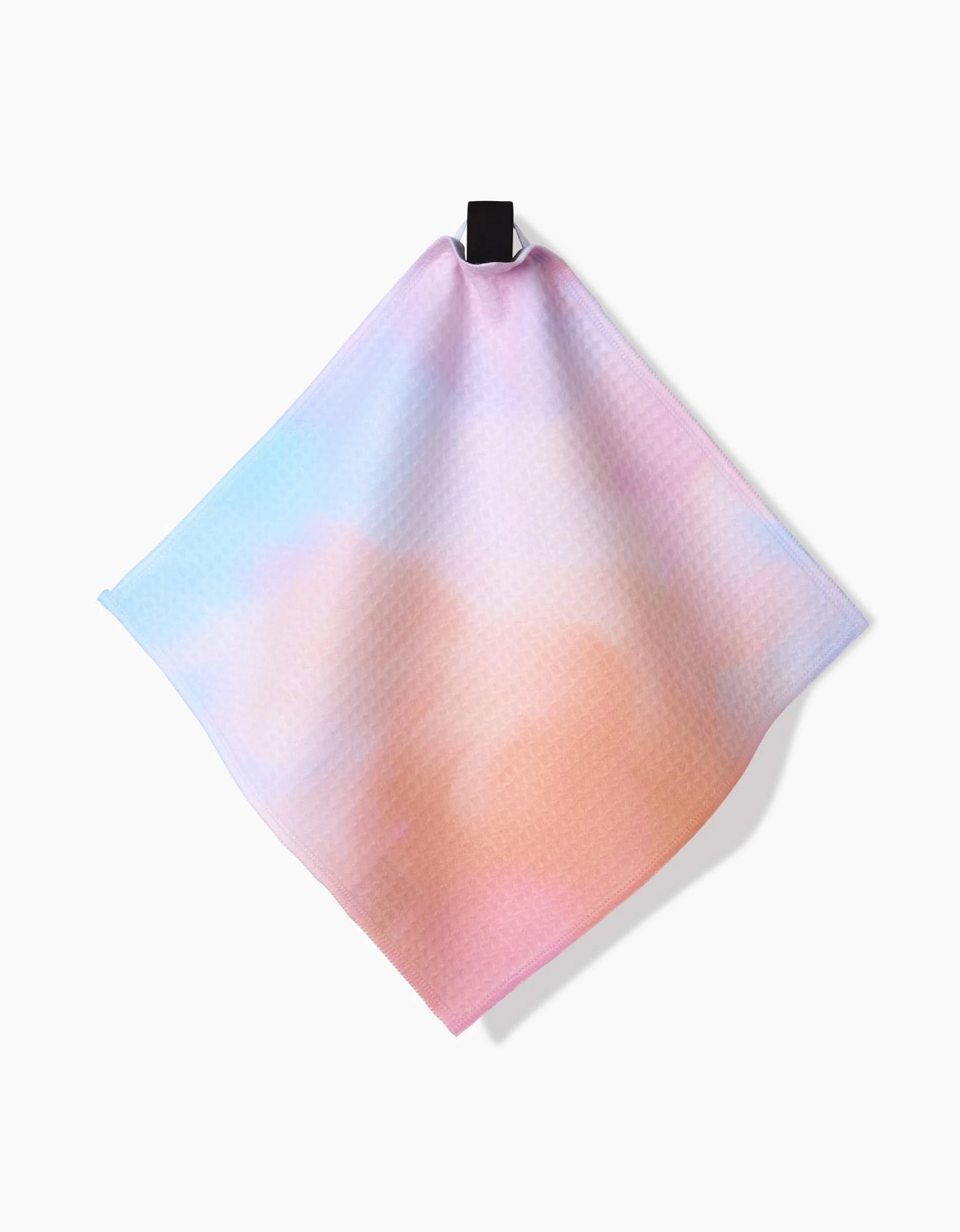 The GEOMETRY Cotton Candy Skies washcloth features soft pastel gradients of pink, orange, and blue, hanging from a black loop against a white background.