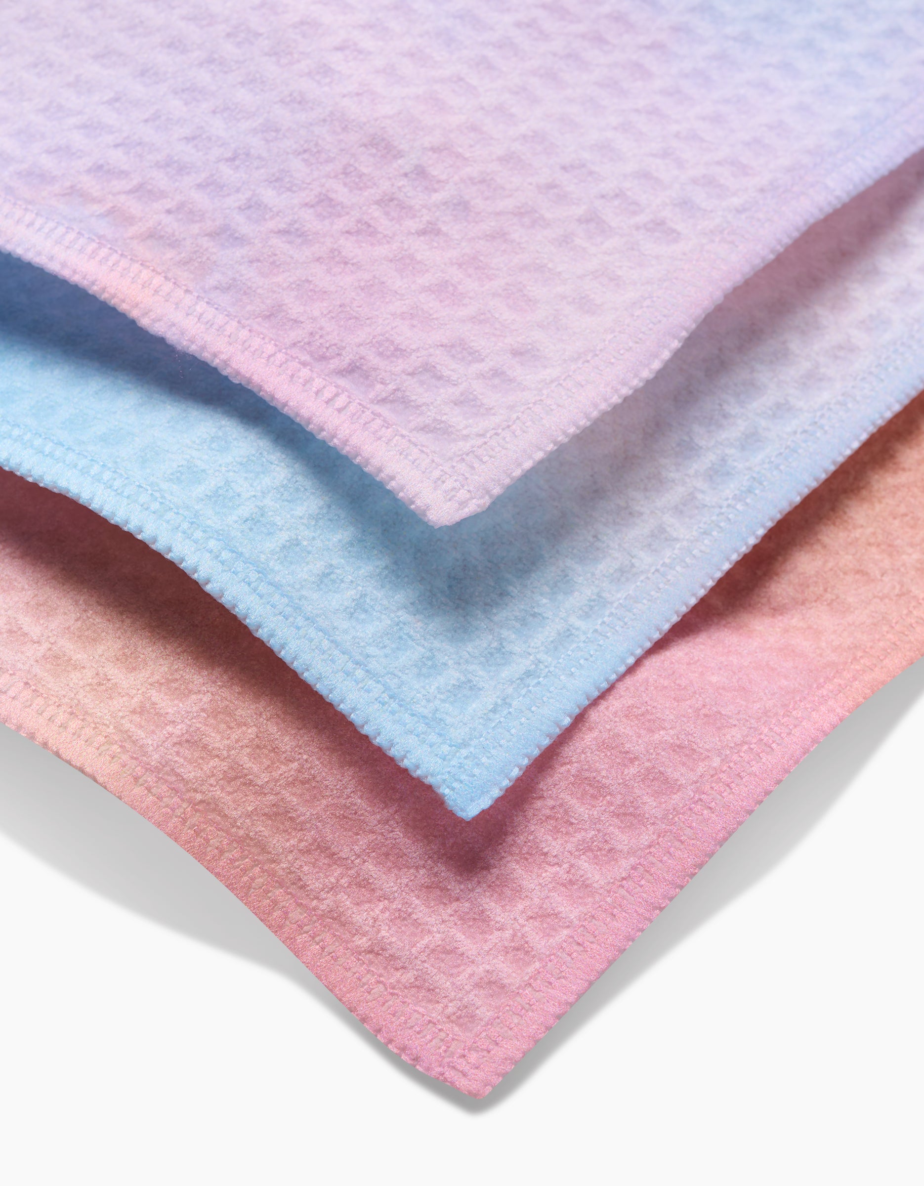 Three ultra-absorbent GEOMETRY Cotton Candy Skies washcloths—lavender, light blue, and light pink with waffle texture and stitched edges—are stacked together, overlapping slightly for a luxe look.
