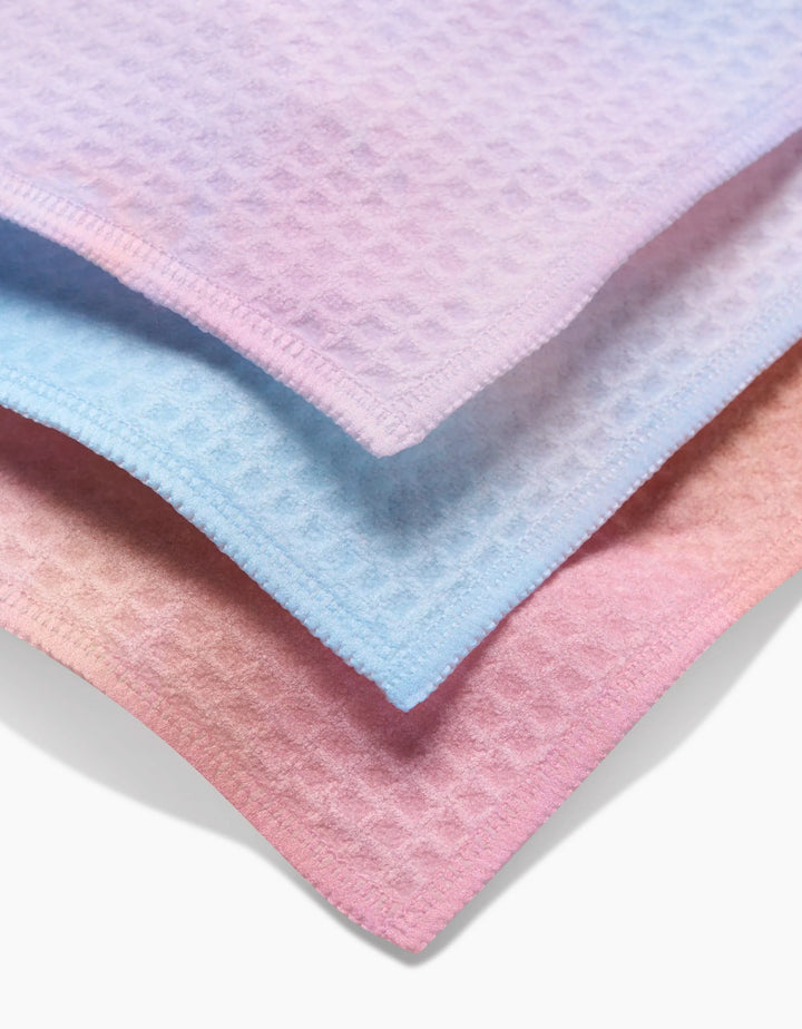 Three ultra-absorbent GEOMETRY Cotton Candy Skies washcloths—lavender, light blue, and light pink with waffle texture and stitched edges—are stacked together, overlapping slightly for a luxe look.