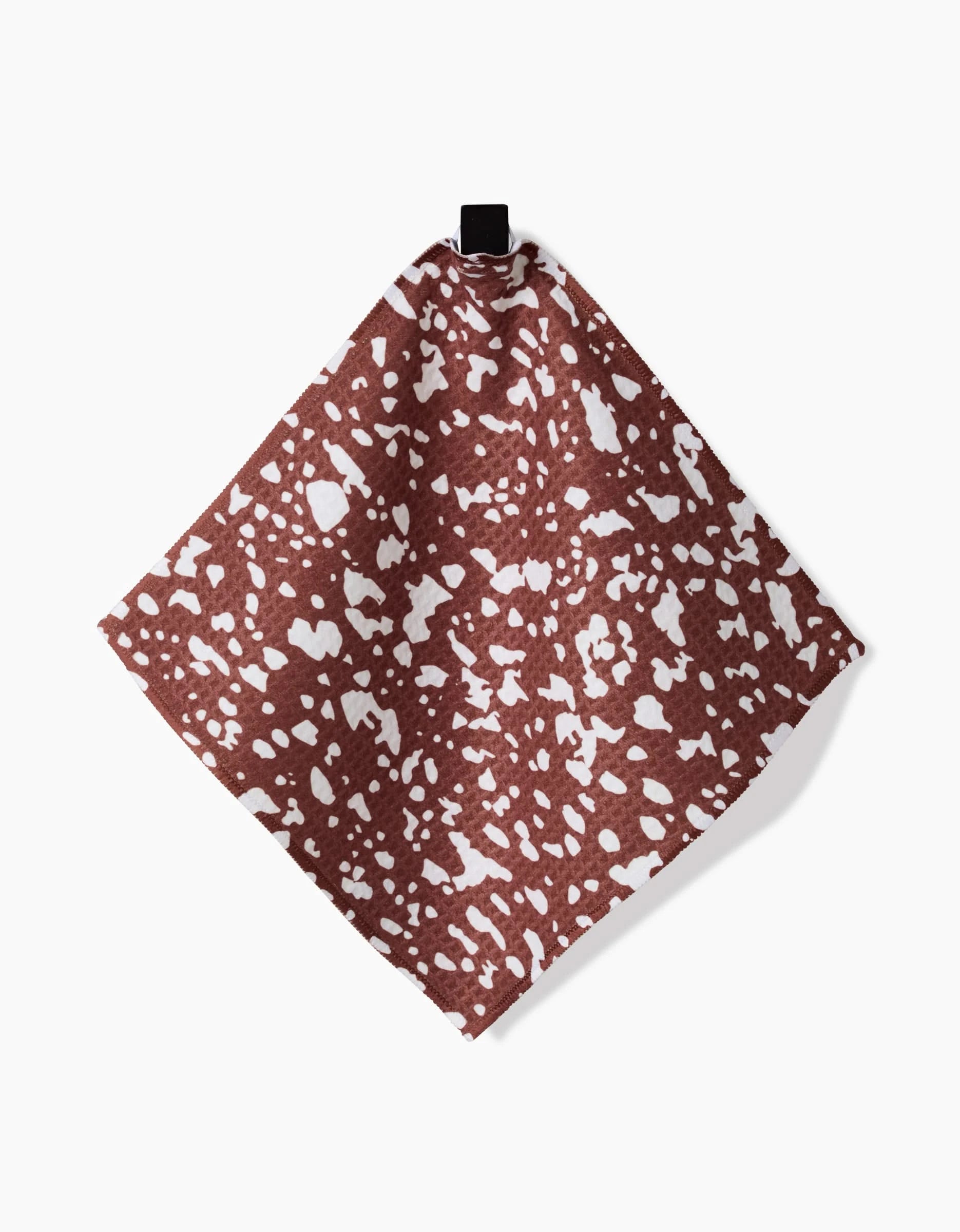 The Cowboy Horse Hide napkin by Hufton Studio features a brown background with an irregular white speckled pattern, made from ultra-absorbent Geoweave technology, and hangs from a small black loop against a plain white surface.