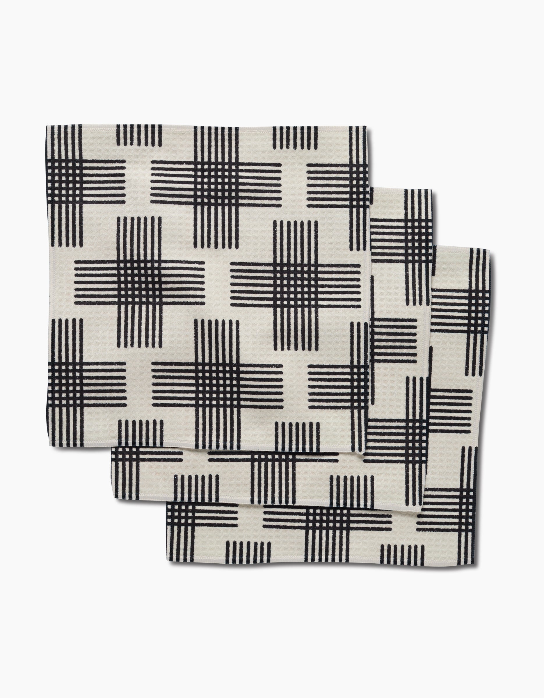 Three folded Cross Hatch napkins by Rosa Kichline, featuring a cream color with black crisscross grid and crafted with Geoweave technology for ultra-absorbency, are stacked slightly overlapping on a white background.