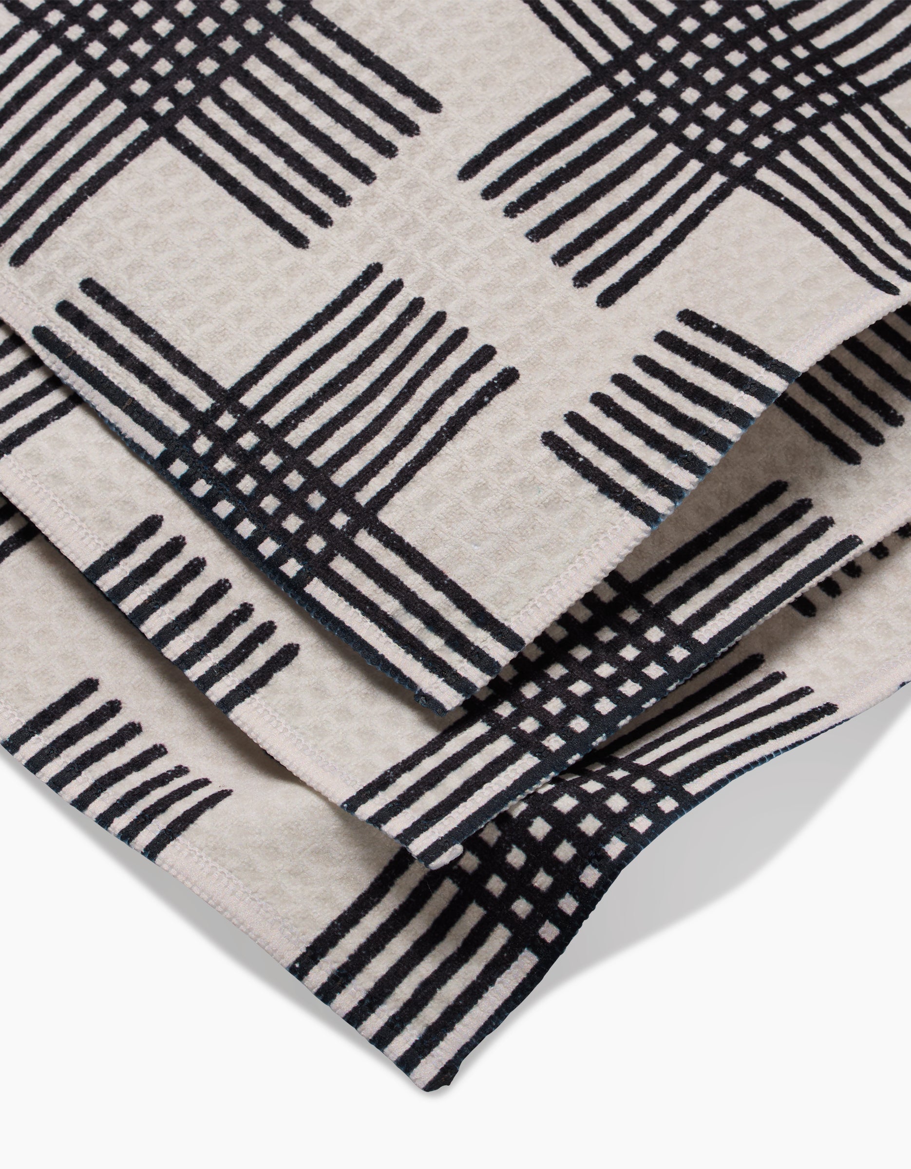 A close-up of folded beige fabric with a bold black crisscross grid, the Cross Hatch Luxe Washcloth by Rosa Kichline features a modern geometric design and ultra-absorbent Geoweave technology.