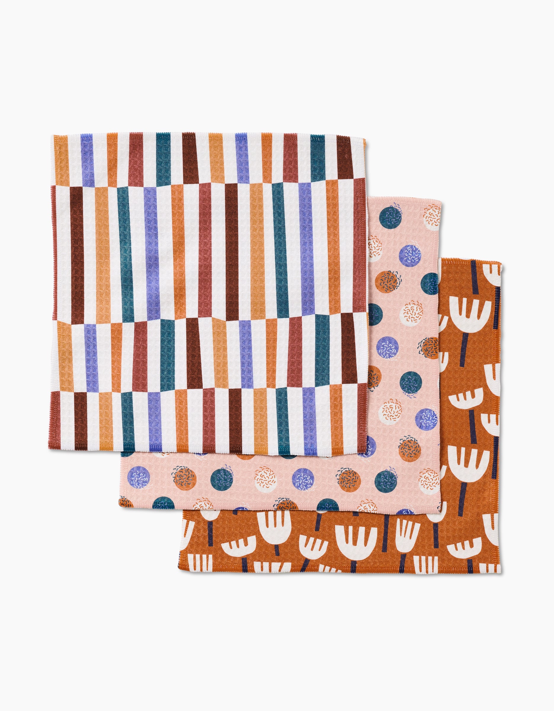 Three folded cloth napkins: one with vertical multicolored stripes, one with blue and orange polka dots on pale fabric, and Claire Iglesias’s Cutout Shapes napkin featuring white abstracts on a rust background.