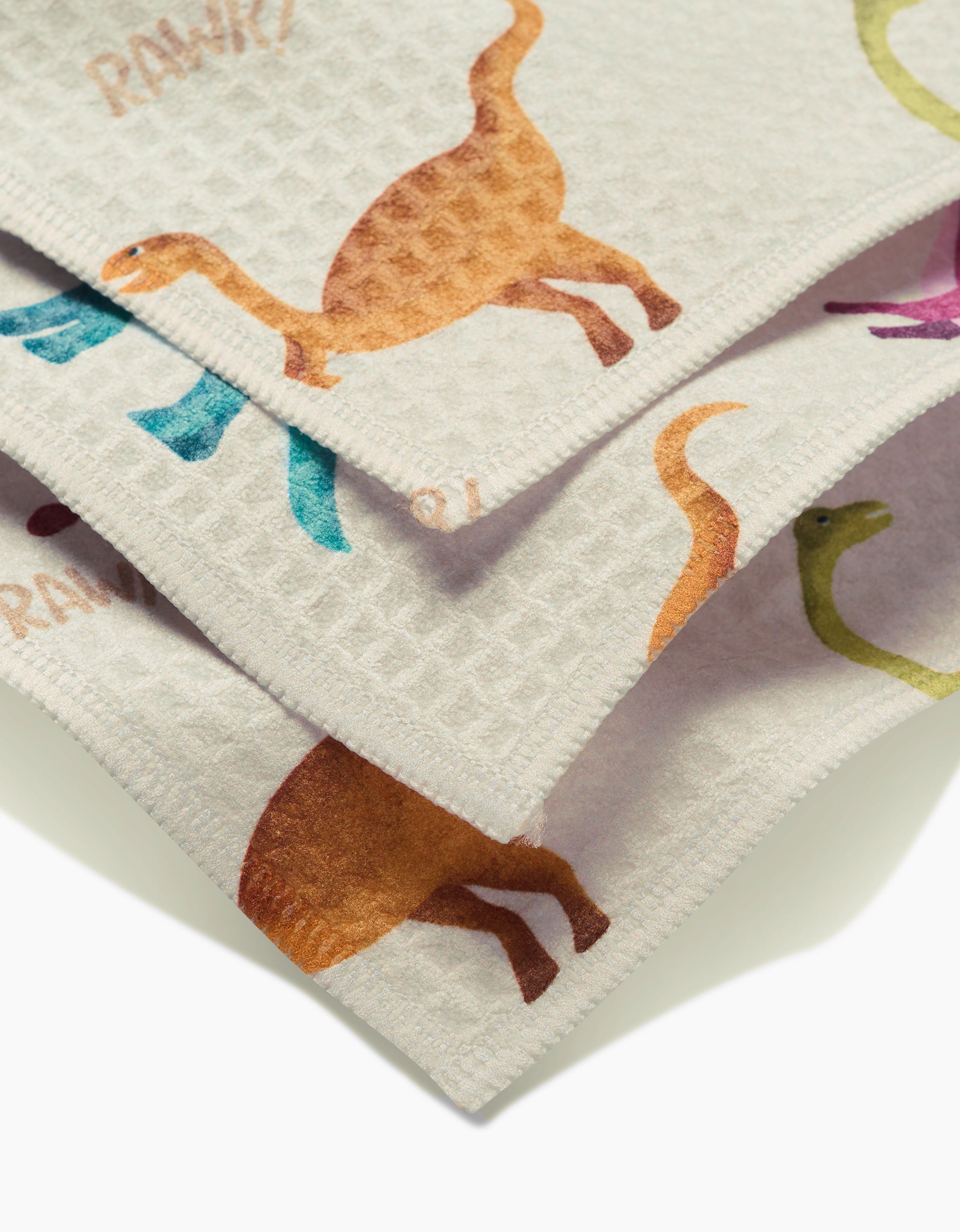 A close-up of three overlapping GEOMETRY Dancing Dinos washcloths, showing colorful long-necked dinosaur prints in orange, green, blue, and purple on light, ultra-absorbent Luxe Washcloth fabric.