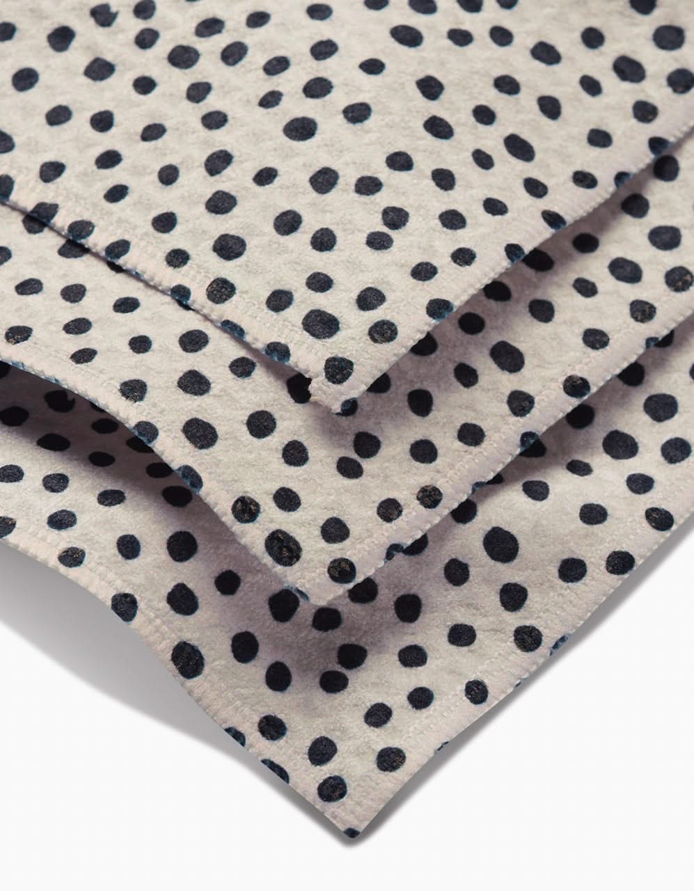 Close-up of GEOMETRYs Dotty Tan: three overlapping sheets of light beige paper with irregular black polka dots and slightly uneven edges, mimicking the ultra-absorbent Luxe Washcloth texture. Dots vary in size and spacing.