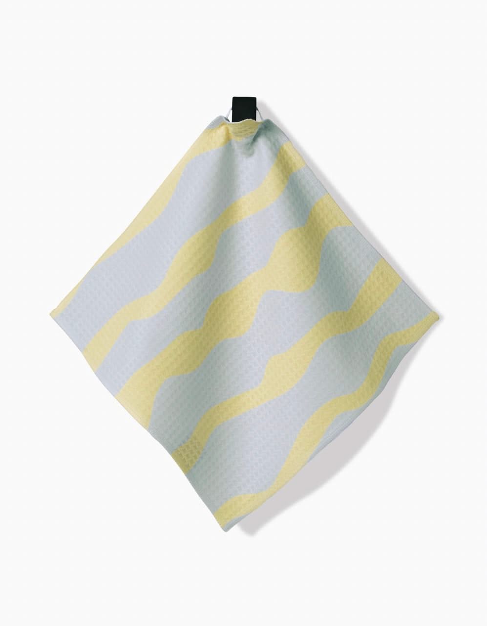 The GEOMETRY Eclipse Twist Washcloth, square-shaped with a subtle textured pattern and wavy yellow and light blue stripes, hangs from a black loop against a plain white background.