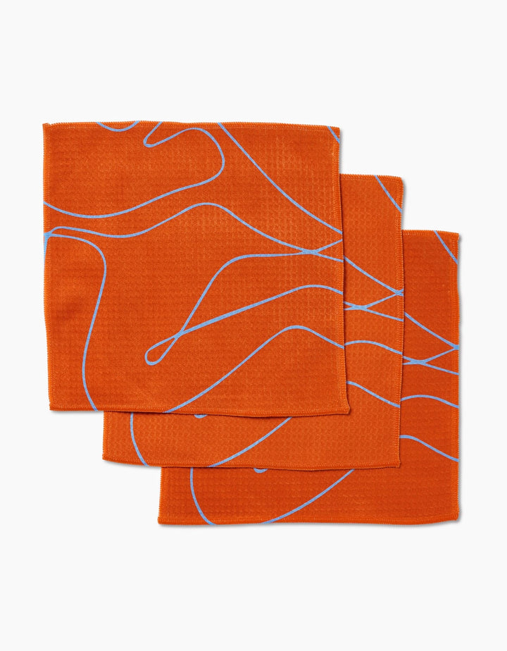 Three Energetic Orange napkins by GEOMETRY with wavy blue lines overlap on a white background. Featuring Geoweave technology, these ultra-absorbent Luxe Washcloths combine style and function for your table.