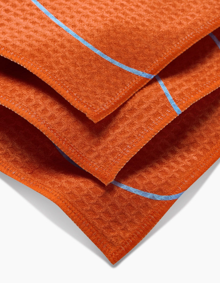 Close-up of three stacked GEOMETRY Energetic Orange washcloths featuring a waffle texture and thin blue grid lines, crafted with ultra-absorbent Geoweave technology.