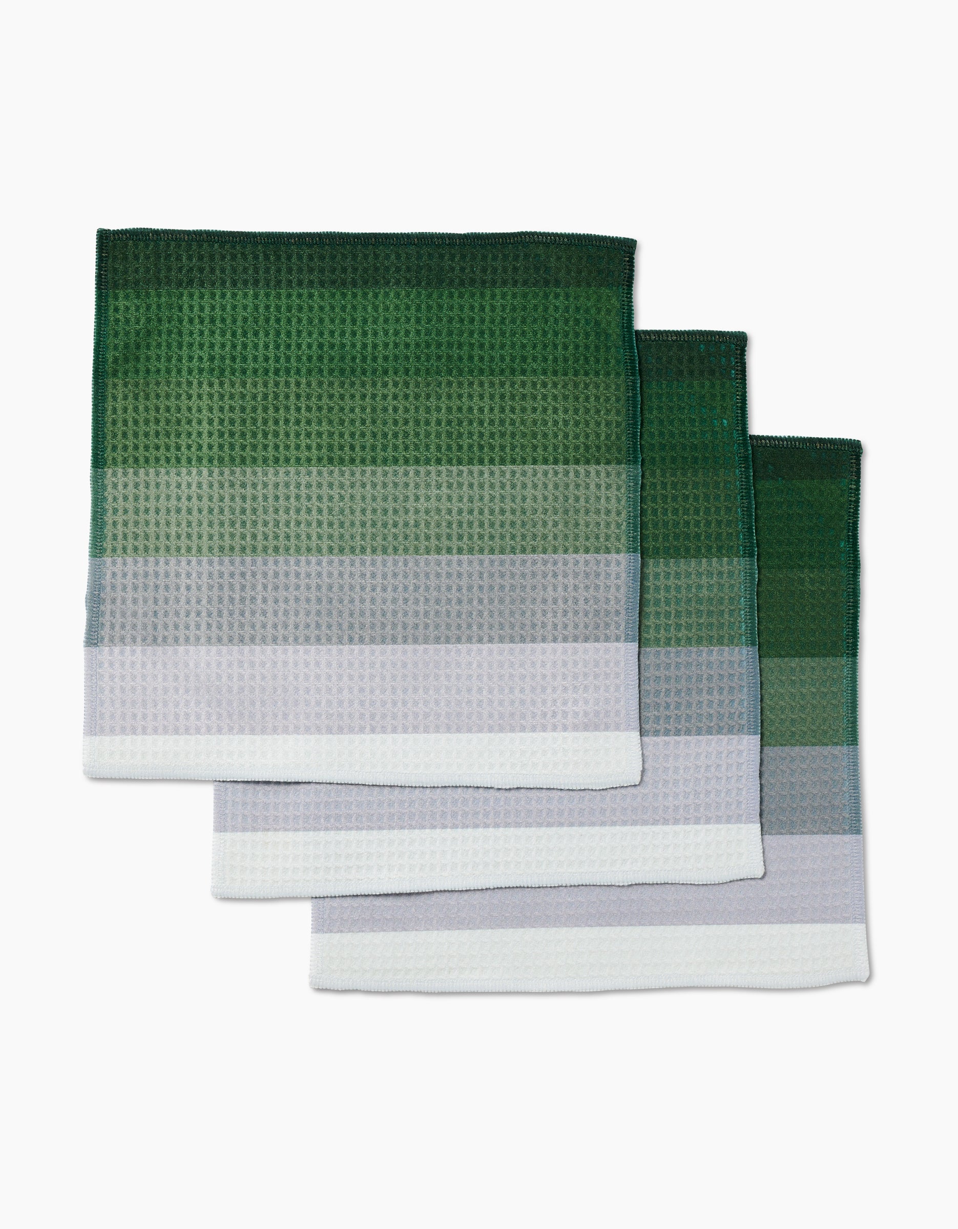 Three Grassy Levels kitchen towels by GEOMETRY, featuring Geoweave technology for ultra-absorbency, have a waffle texture and gradient design transitioning from dark green to light green to white. Towels are shown in a staggered stack.