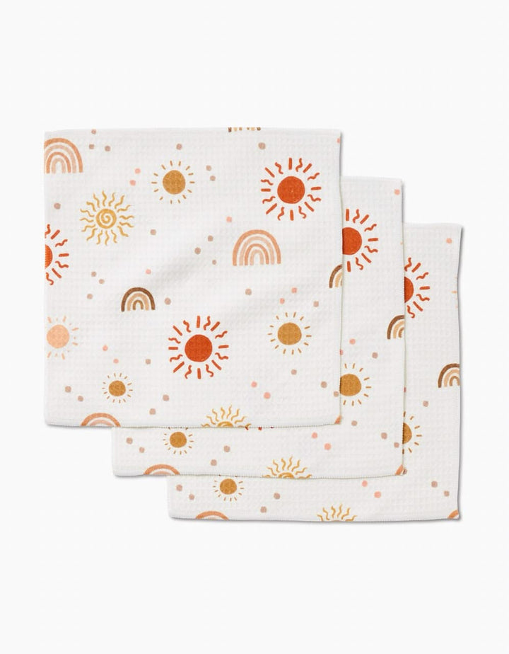 Three ultra-absorbent GEOMETRY Happy Skies white cloth napkins, featuring a repeating sun, rainbow, and dot pattern in orange, yellow, and brown tones, are stacked slightly offset on a white background.