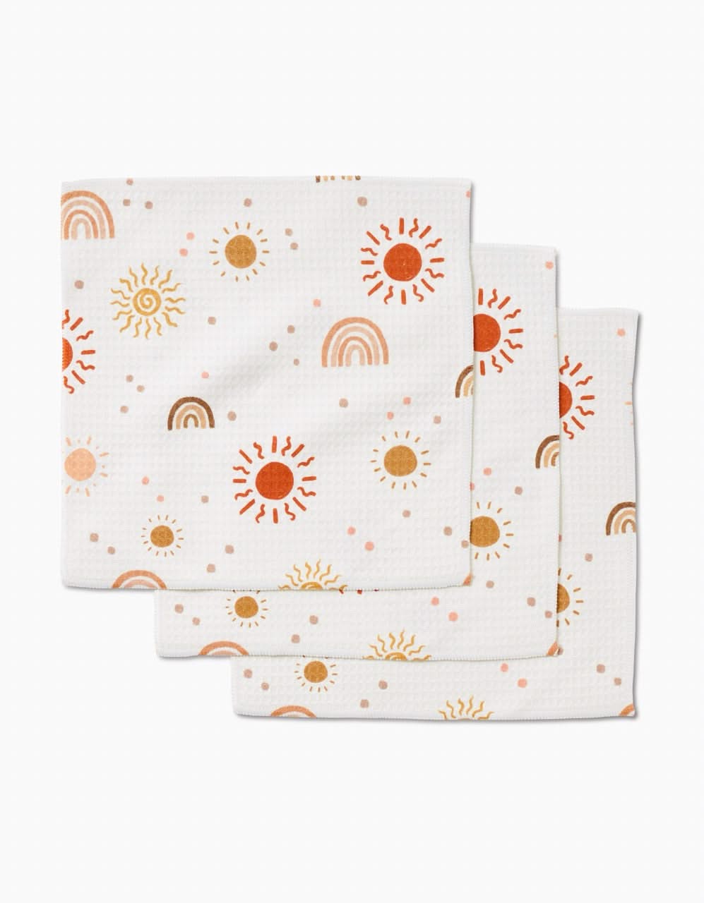 Three ultra-absorbent GEOMETRY Happy Skies white cloth napkins, featuring a repeating sun, rainbow, and dot pattern in orange, yellow, and brown tones, are stacked slightly offset on a white background.