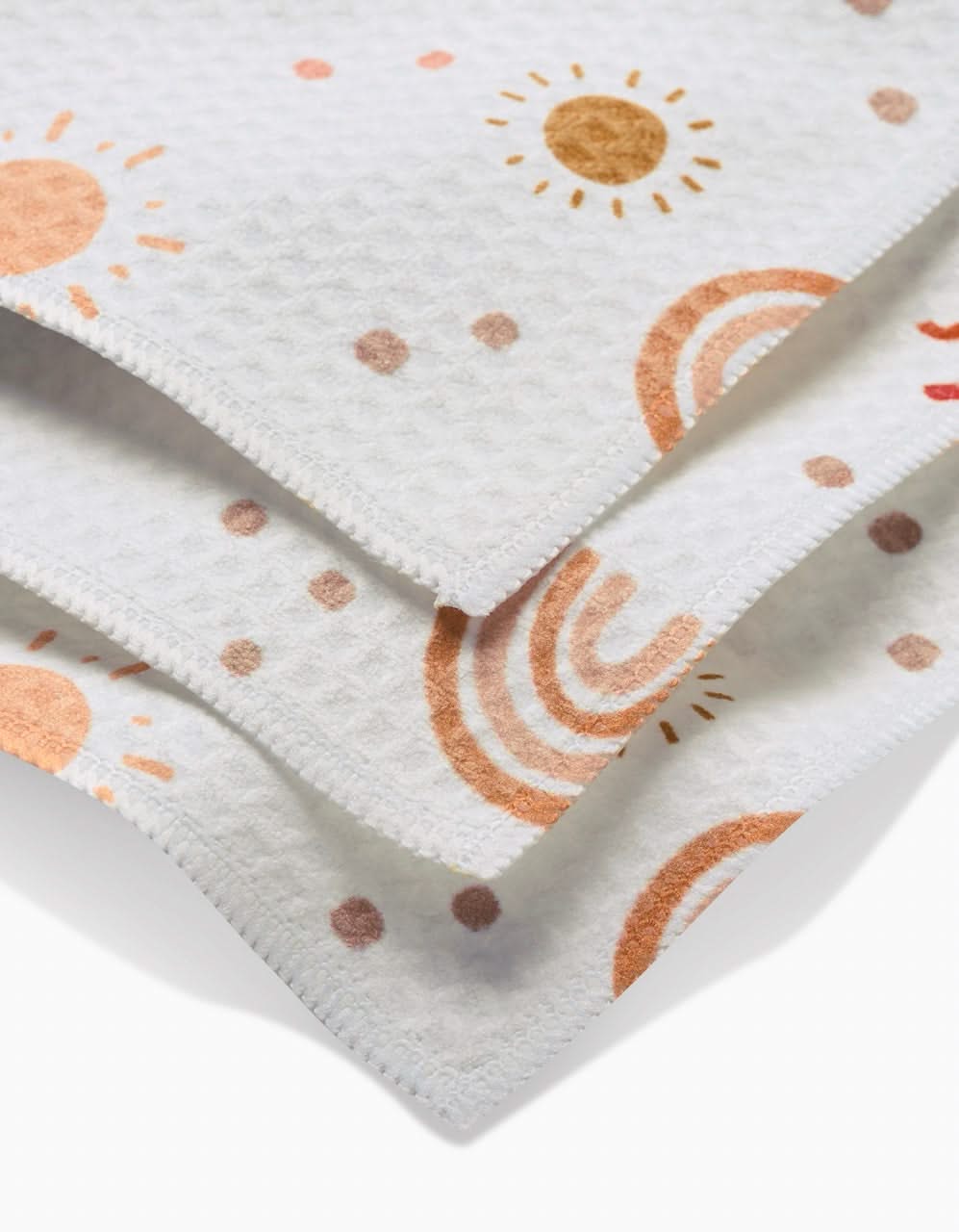 A stack of GEOMETRY Luxe Washcloths in the Happy Skies print displays ultra-absorbent white textured fabric with colorful suns, rainbows, and dots in shades of orange, brown, and peach.