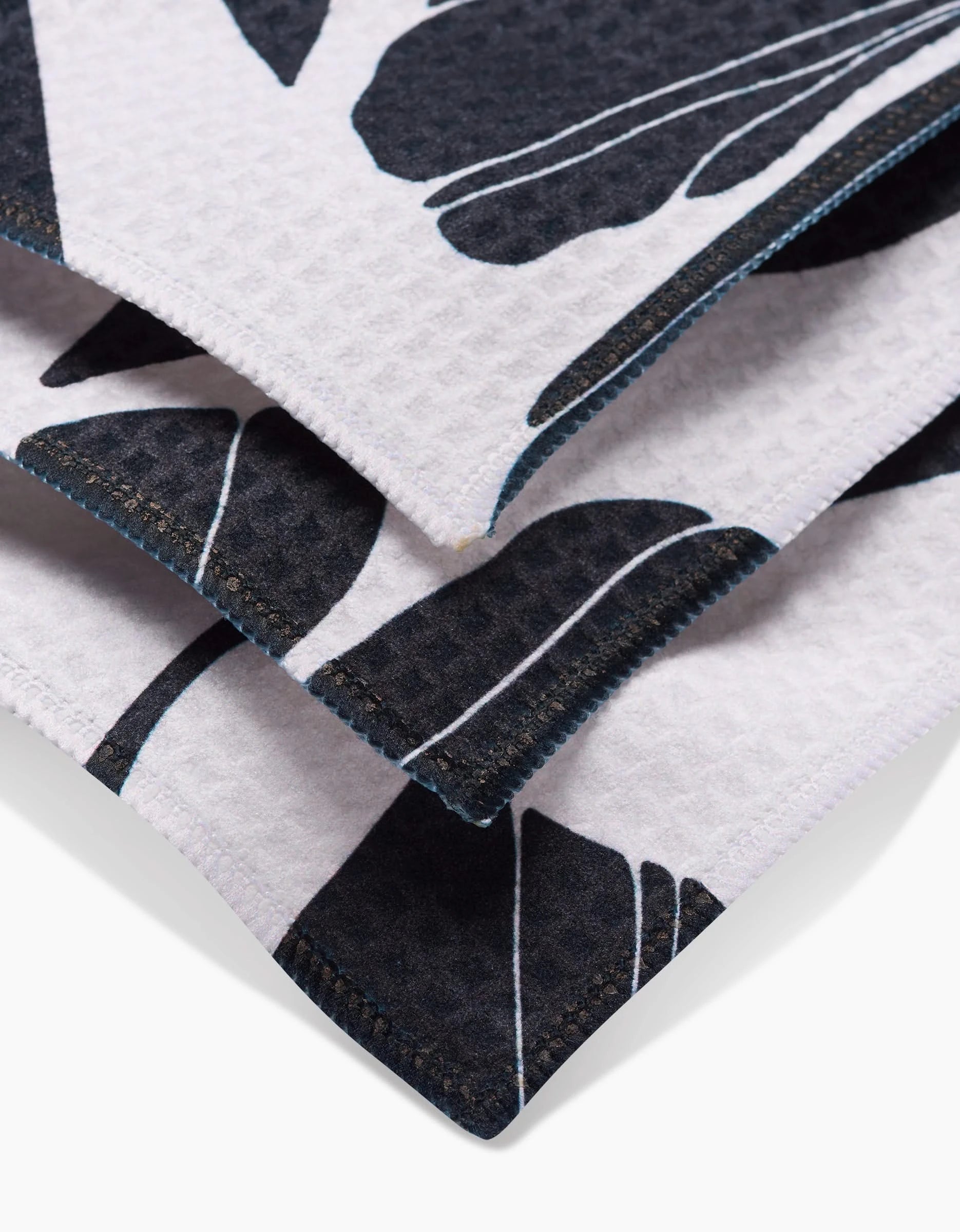 Close-up of the Harmony washcloth by GEOMETRY, made from recycled materials, featuring folded white textured fabric with a black leaf pattern and dark blue stitched edges.