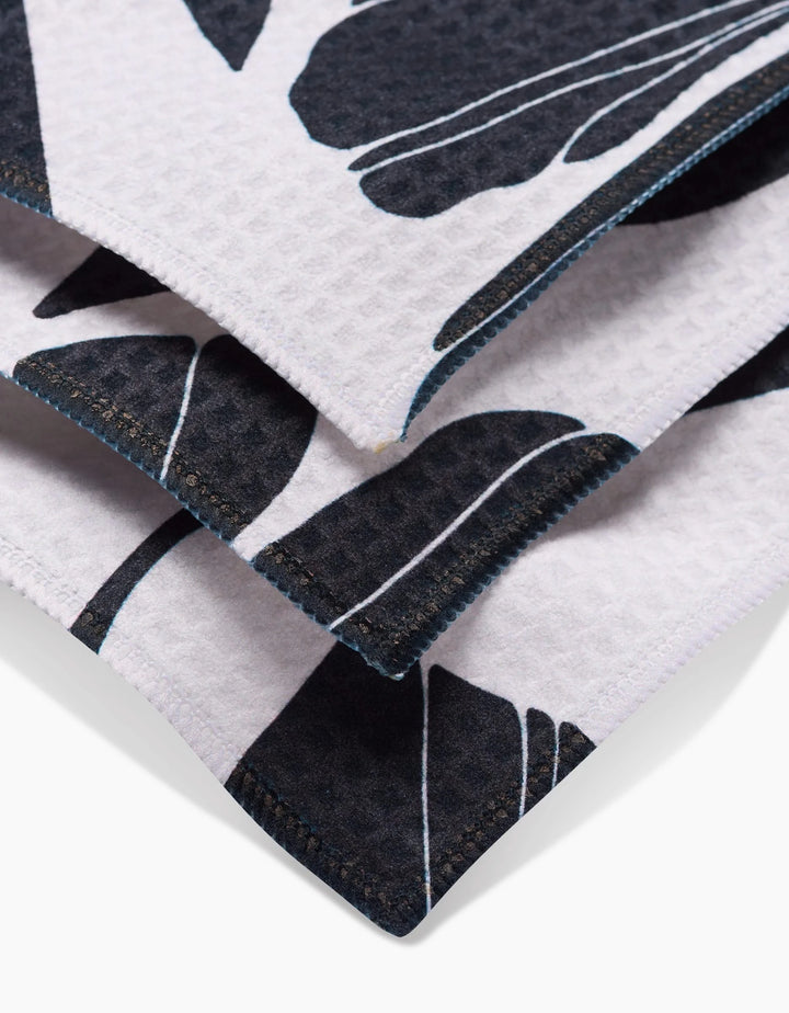 Close-up of the Harmony washcloth by GEOMETRY, made from recycled materials, featuring folded white textured fabric with a black leaf pattern and dark blue stitched edges.