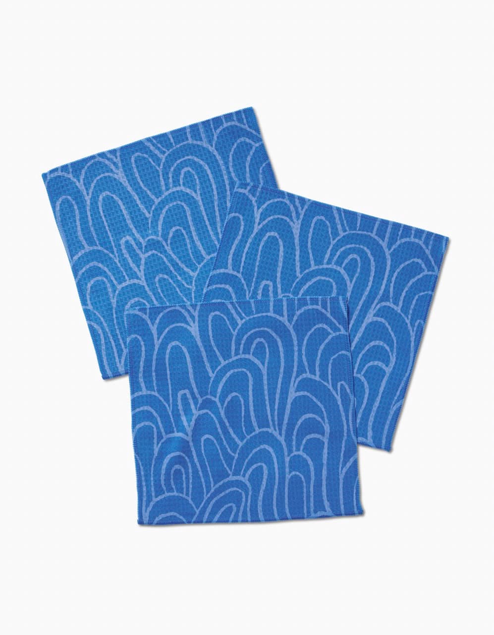 Three High Tide napkins by Sareka Unique, featuring a wavy blue abstract pattern in varied shades and crafted with ultra-absorbent Geoweave technology, are overlapped on a white background.