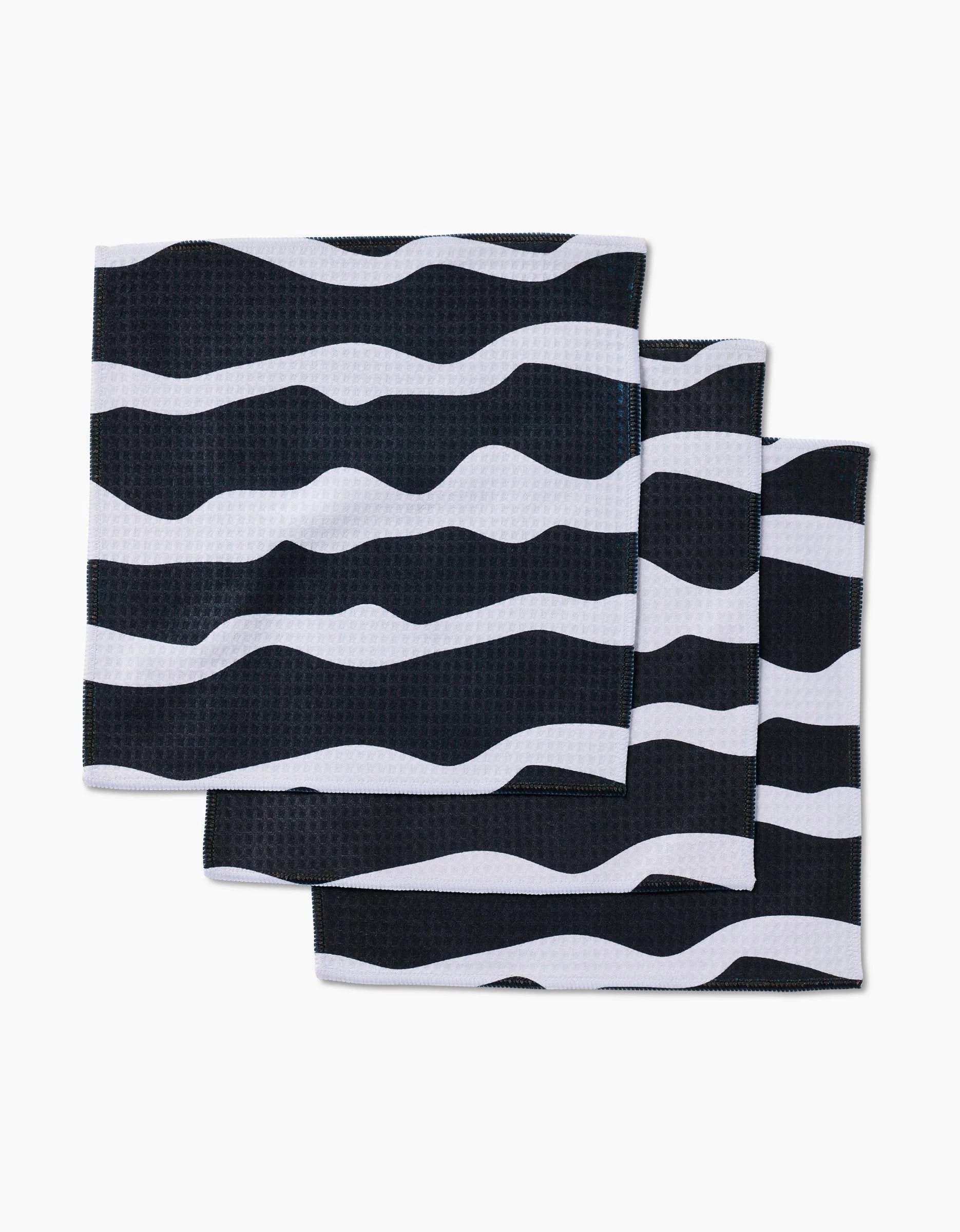 Three GEOMETRY Ink Twist black and white cloth napkins made from recycled materials, featuring a wavy striped pattern, are stacked unevenly against a white background.