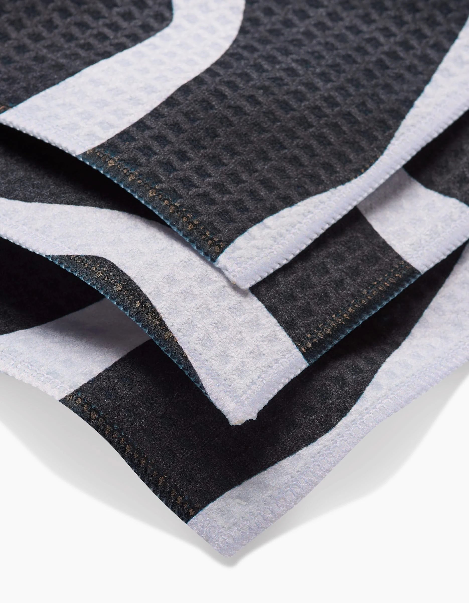 Close-up of three overlapping black and white GEOMETRY Ink Twist Luxe Washcloths with a waffle texture, made from ultra-absorbent recycled materials, showcasing stitched edges and a geometric pattern.