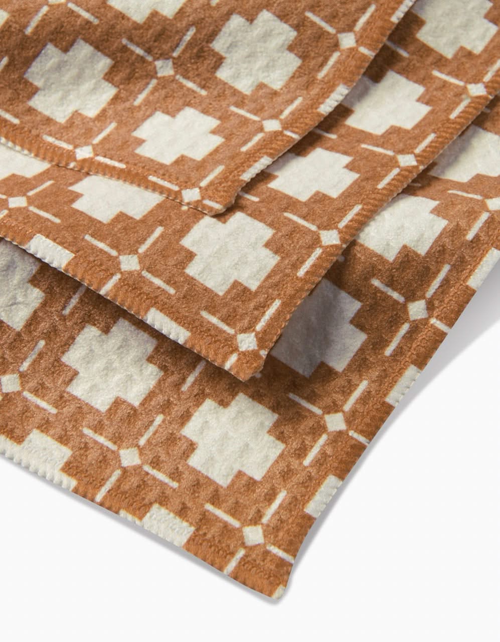 Close-up of the Jackson blanket by Charlie Rowan Designs, showcasing a soft, cozy fabric with white and brown geometric diamond and plus motifs. Made from recycled materials and finished with neatly stitched edges.