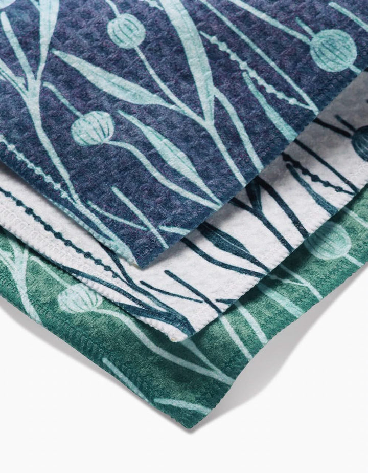 Three folded Kelp washcloths by Katie OShea, in blue, white, and green, each with a botanical leaf and flower bud pattern. Ultra-absorbent and neatly stacked, the towels are viewed from above.