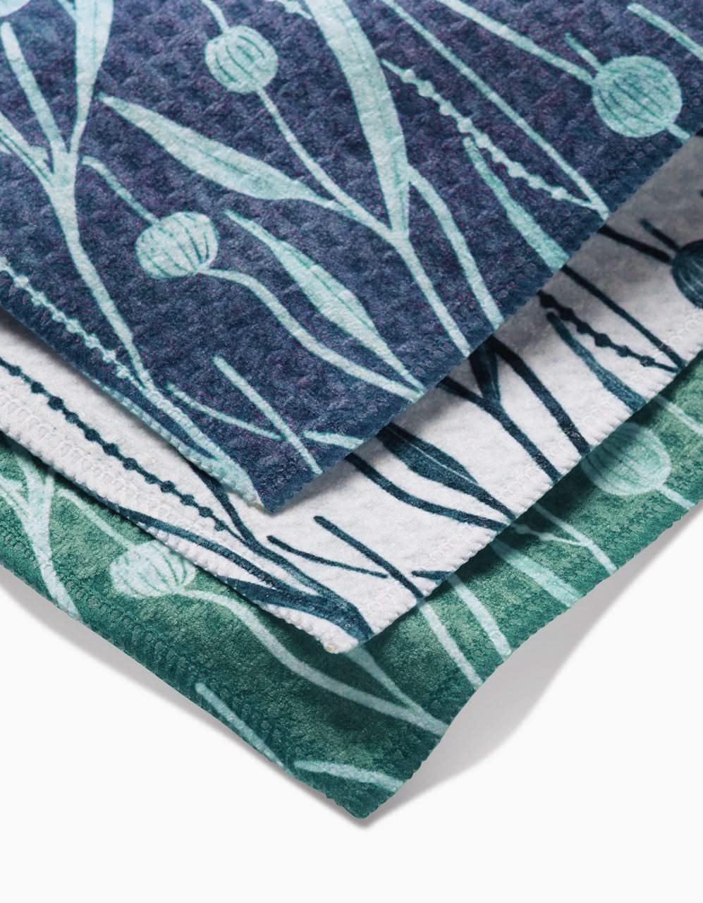 Three folded Kelp washcloths by Katie OShea, in blue, white, and green, each with a botanical leaf and flower bud pattern. Ultra-absorbent and neatly stacked, the towels are viewed from above.