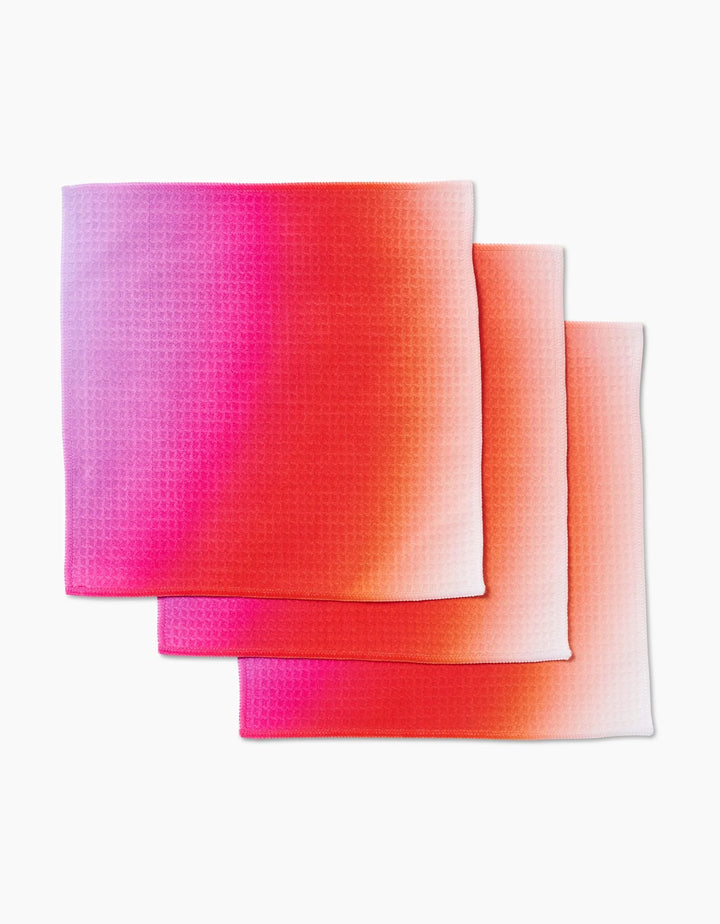 Three folded GEOMETRY Lively Hue washcloths are stacked, each displaying a gradient of pink, red, orange, and white on a textured surface with ultra-absorbent Geoweave technology.