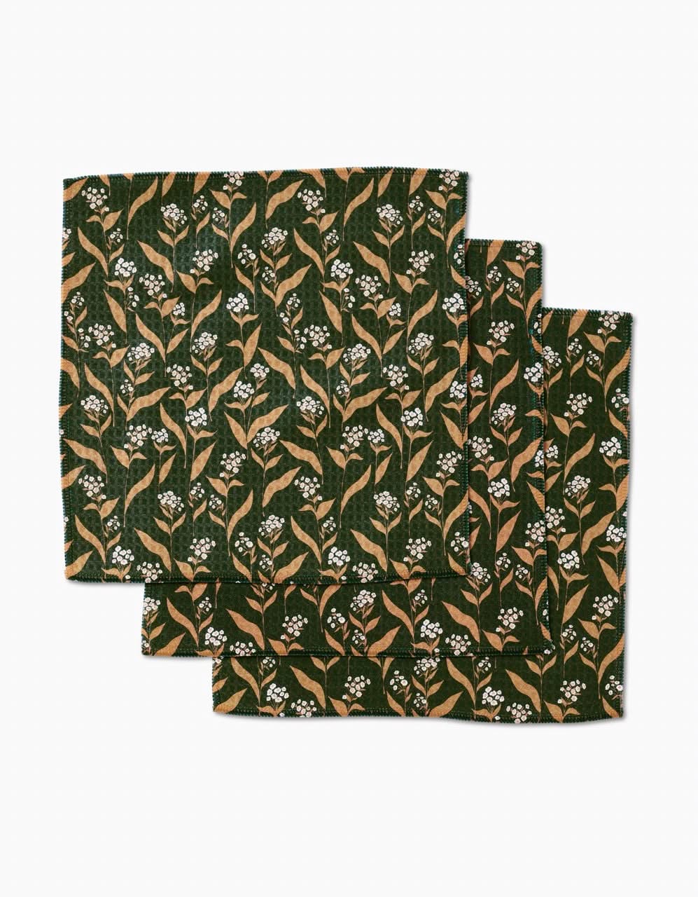 The Loretta washcloth set by Charlie Rowan Designs features three folded napkins in dark green with a tan leaf and white flower pattern, crafted from recycled materials for an eco-friendly touch.
