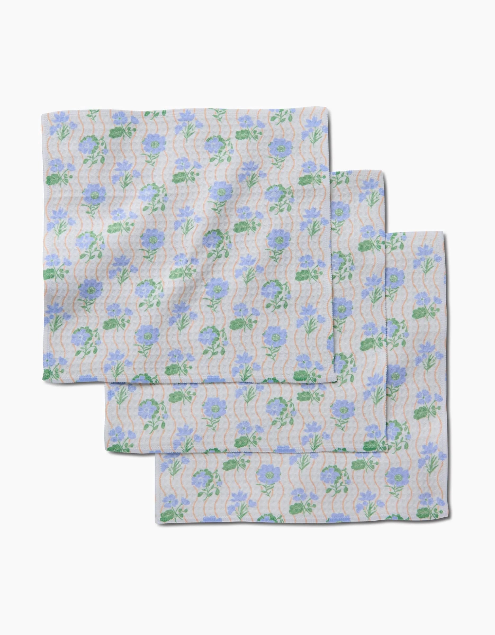 Three neatly folded Danelys Sidron Morning Wave washcloths, featuring blue flowers, green leaves, and wavy lines on a light background, display their ultra-absorbent Geoweave technology on a white surface.