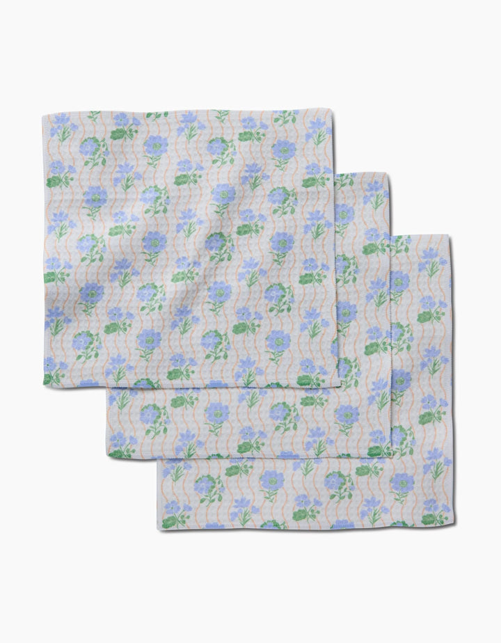 Three neatly folded Danelys Sidron Morning Wave washcloths, featuring blue flowers, green leaves, and wavy lines on a light background, display their ultra-absorbent Geoweave technology on a white surface.