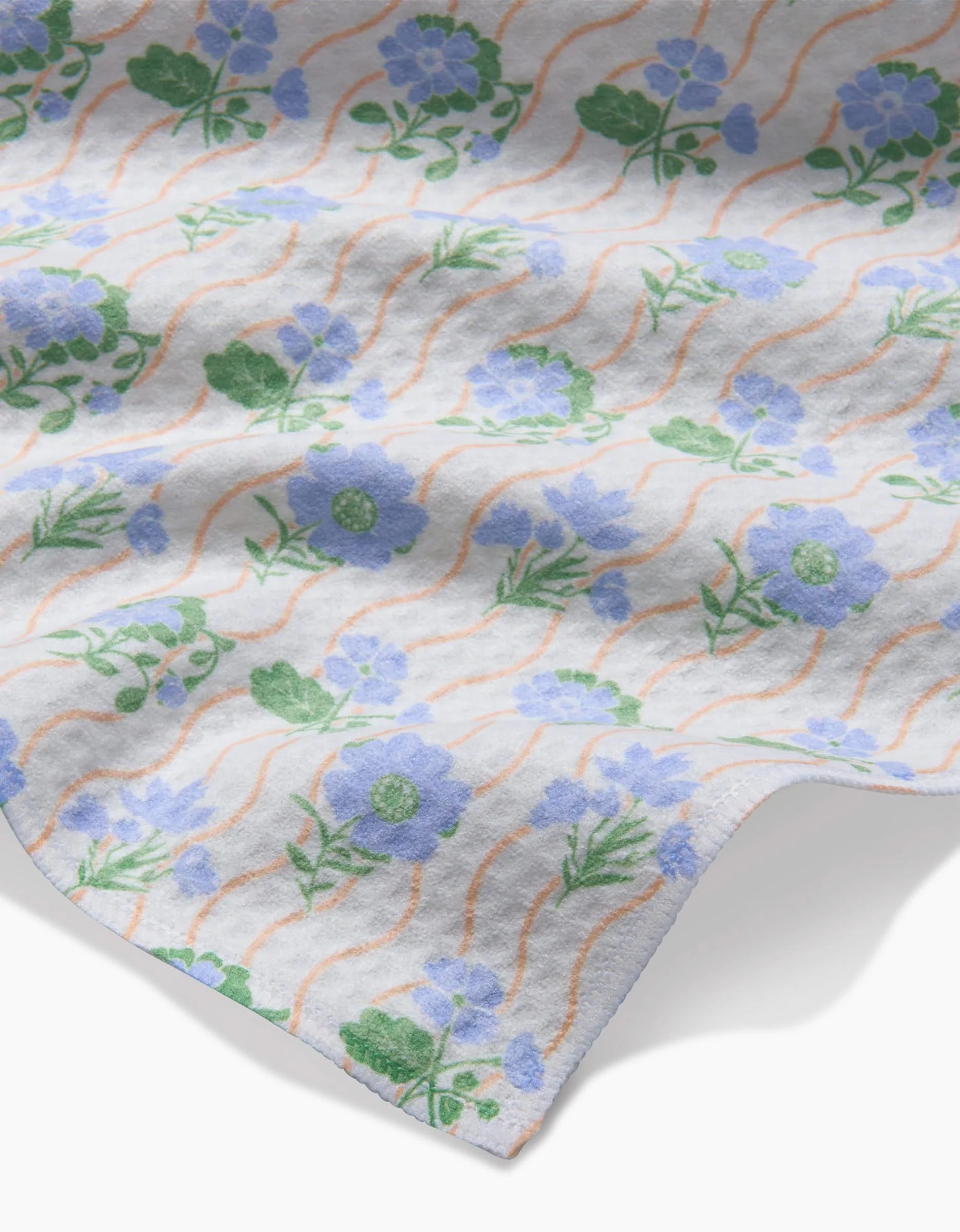 A close-up of Danelys Sidrons Morning Wave Luxe Washcloth shows soft Geoweave fabric with blue and green floral patterns and wavy orange lines; its ultra-absorbent edge is slightly curled.