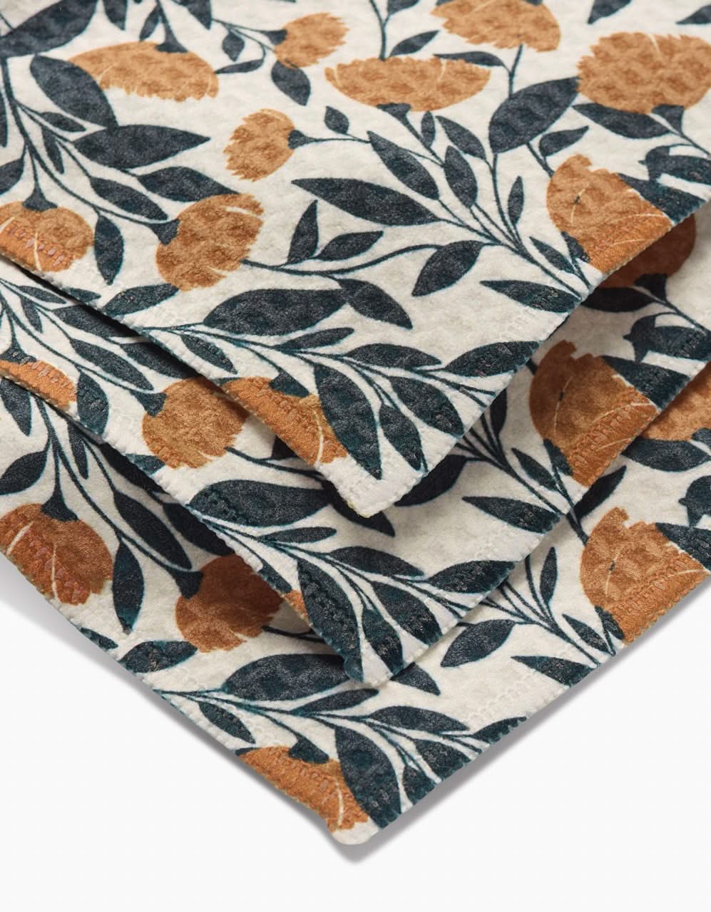 A close-up of the Naomi washcloth by Charlie Rowan Designs, made from recycled materials, with orange-brown flowers and dark green leaves on a light cream background.
