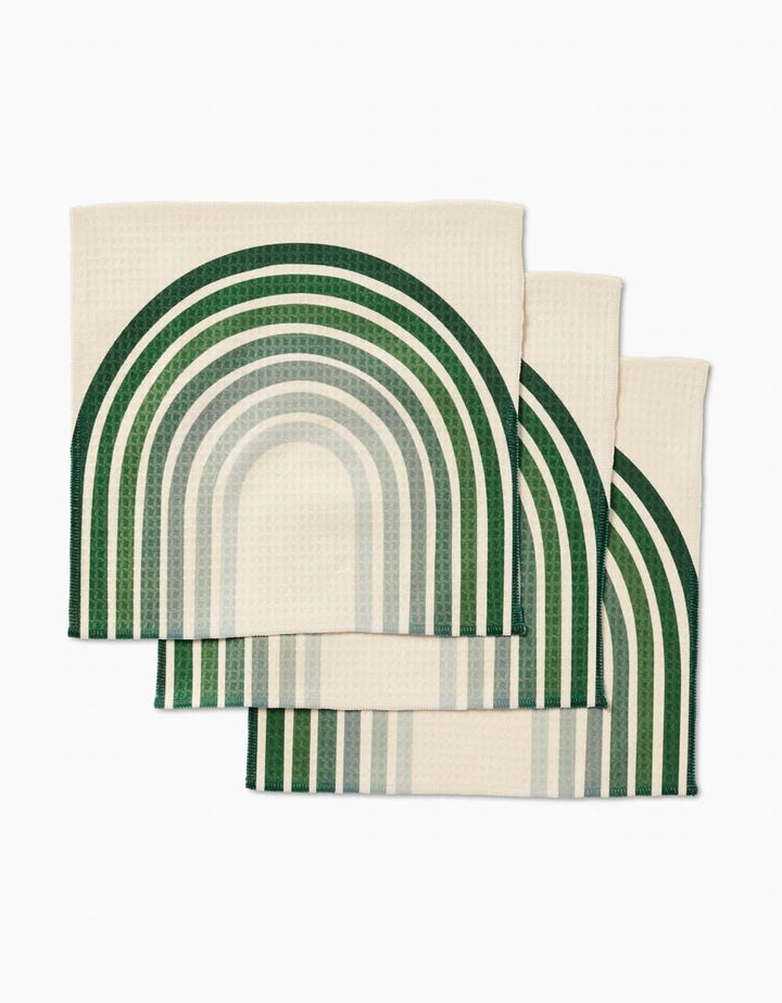 Three cream-colored GEOMETRY Olive Arches Luxe Washcloths, featuring a green and teal geometric rainbow pattern, are neatly stacked with overlapping curved designs against a white background.