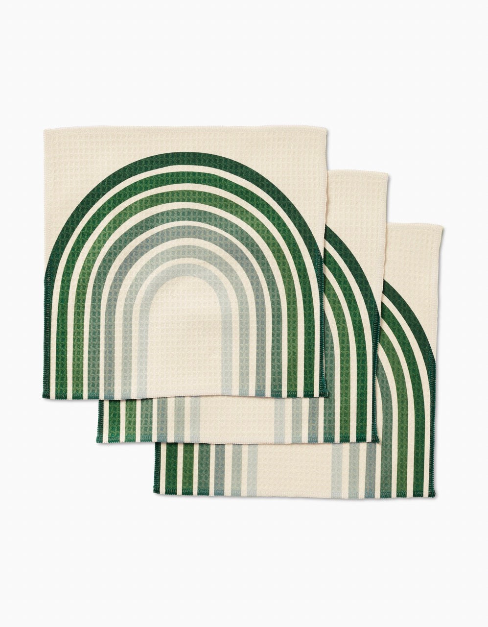 Three cream-colored GEOMETRY Olive Arches Luxe Washcloths, featuring a green and teal geometric rainbow pattern, are neatly stacked with overlapping curved designs against a white background.