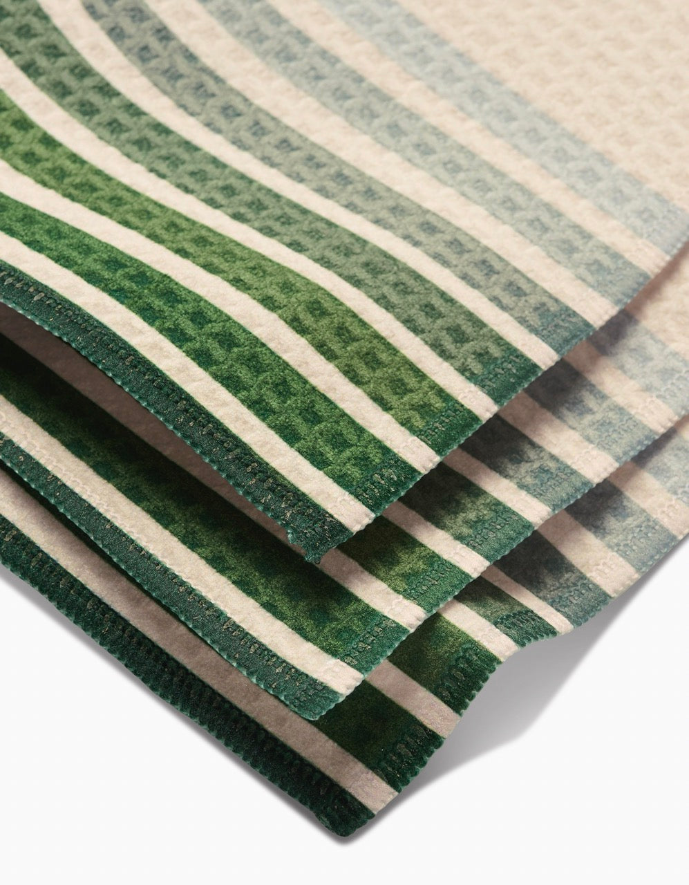 A close-up of folded GEOMETRY Olive Arches washcloths, featuring green and white gradient stripes, a textured pattern, and green-stitched edges that highlight their ultra-absorbent Geoweave technology.
