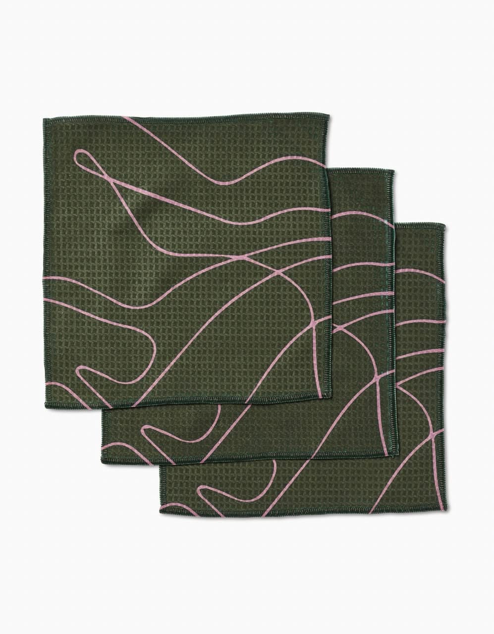 Three Olive Martini Luxe Washcloths by GEOMETRY, featuring a dark green textured surface with abstract pink lines and innovative Geoweave technology, are stacked with slight overlap on a white background.
