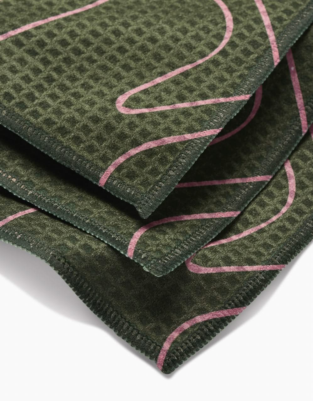 A close-up of folded, textured, dark green fabric with a waffle weave pattern and pink curved lines, featuring GEOMETRY’s ultra-absorbent Luxe Washcloths in Olive Martini made with Geoweave technology.