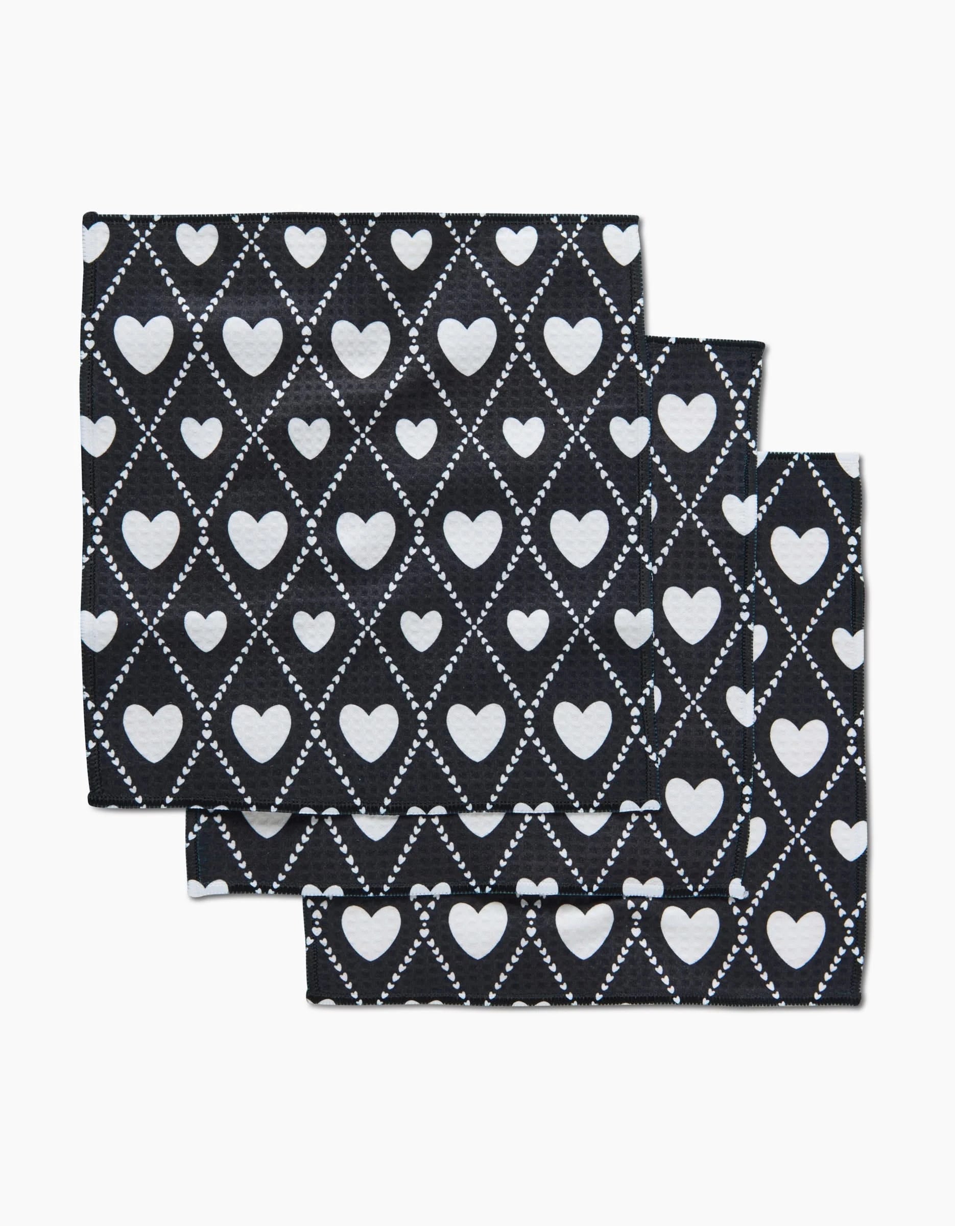 Three folded Onyx Love Lattice washcloths by Charlie Rowan Designs, featuring a black backdrop with white hearts in diamond patterns outlined by dotted lines, are slightly overlapped on a white background.