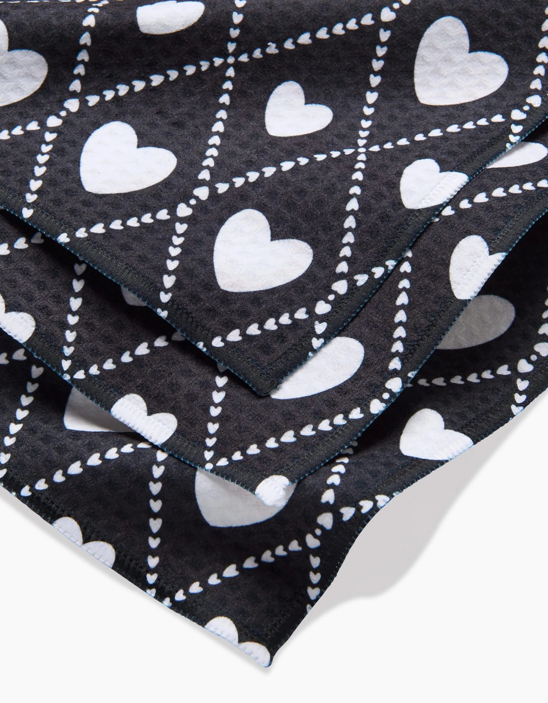 A close-up of Charlie Rowan Designs Onyx Love Lattice Luxe Washcloths, featuring diagonal rows of large and small white hearts on black fabric, crafted with ultra-absorbent Geoweave technology for both style and performance.