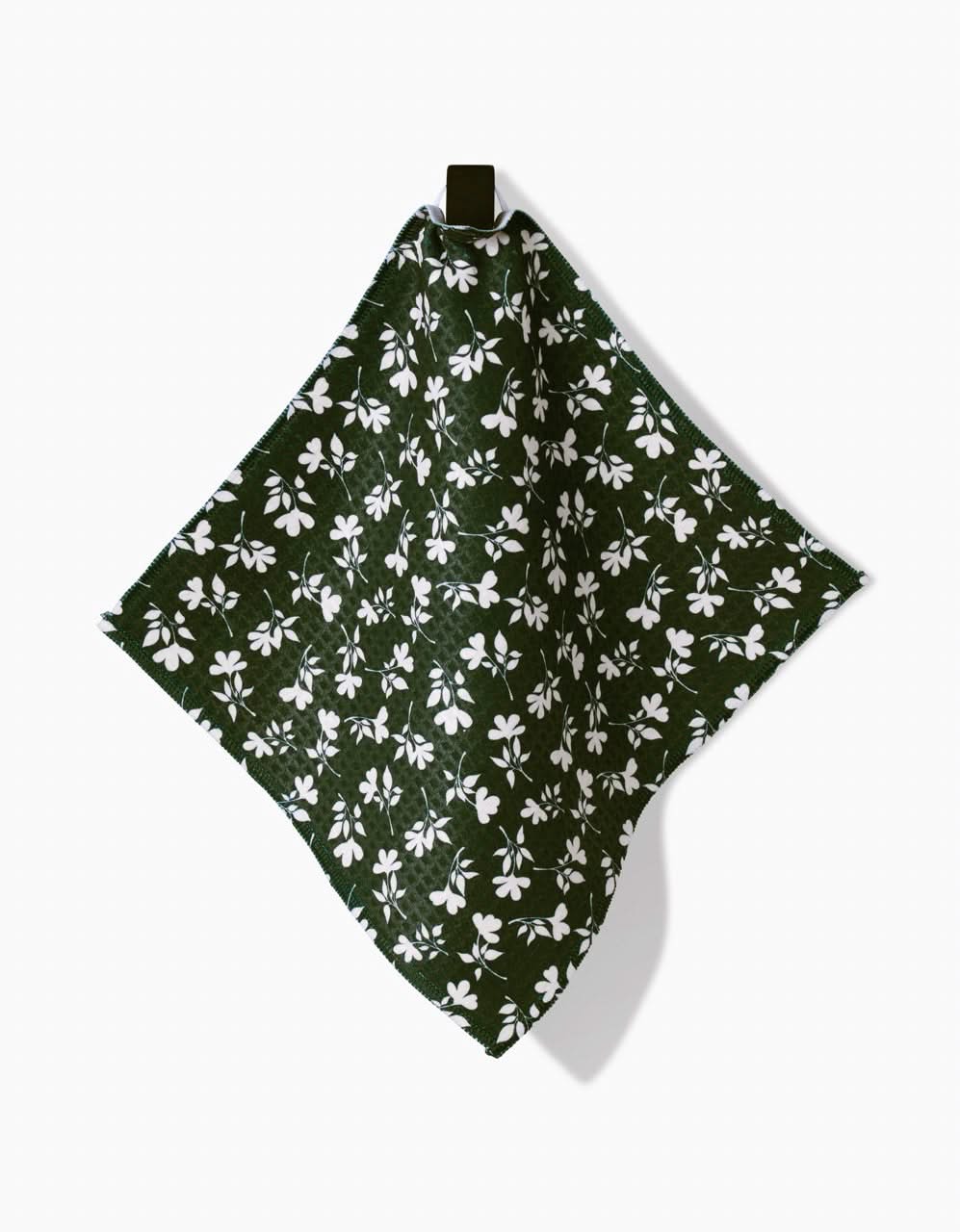 The Penelope Luxe Washcloth by Charlie Rowan Designs, featuring a white floral pattern on dark green and made with ultra-absorbent Geoweave technology, hangs from a black hook against a plain white background.