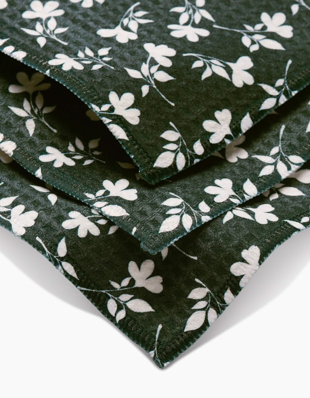 A close-up of stacked Penelope washcloths by Charlie Rowan Designs, featuring dark green fabric with a white floral and vine pattern, scalloped edges, and crafted with Geoweave technology.