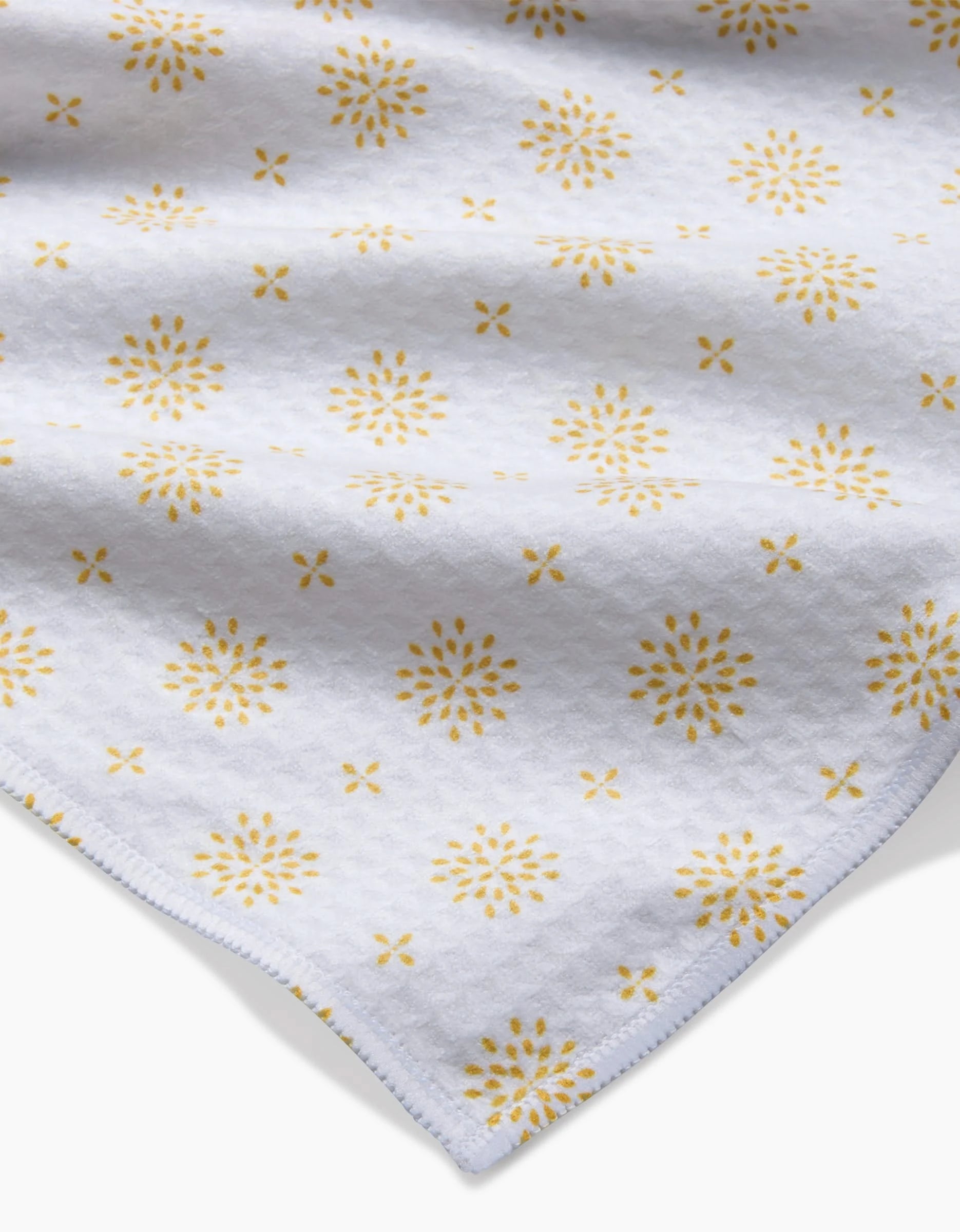 The Spring Morning washcloth by Danelys Sidron features ultra-absorbent Geoweave technology and is crafted from white fabric with a subtle texture, accented by evenly spaced yellow floral motifs. Shown angled to highlight a corner.